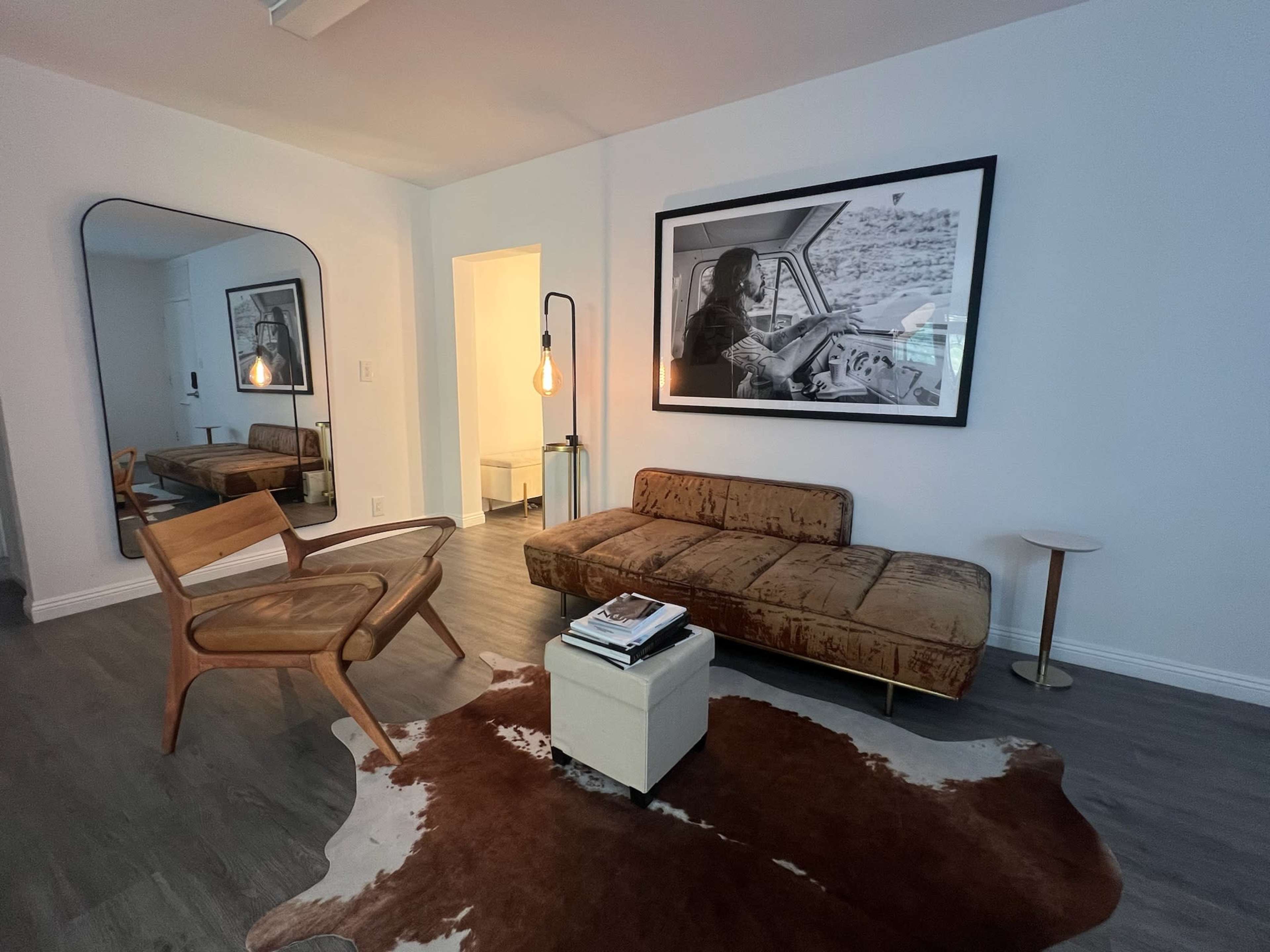 The image shows a modern living room with a brown couch, a wooden chair, a round side table, and a large black-and-white framed photo on the wall, all placed over a cowhide rug.