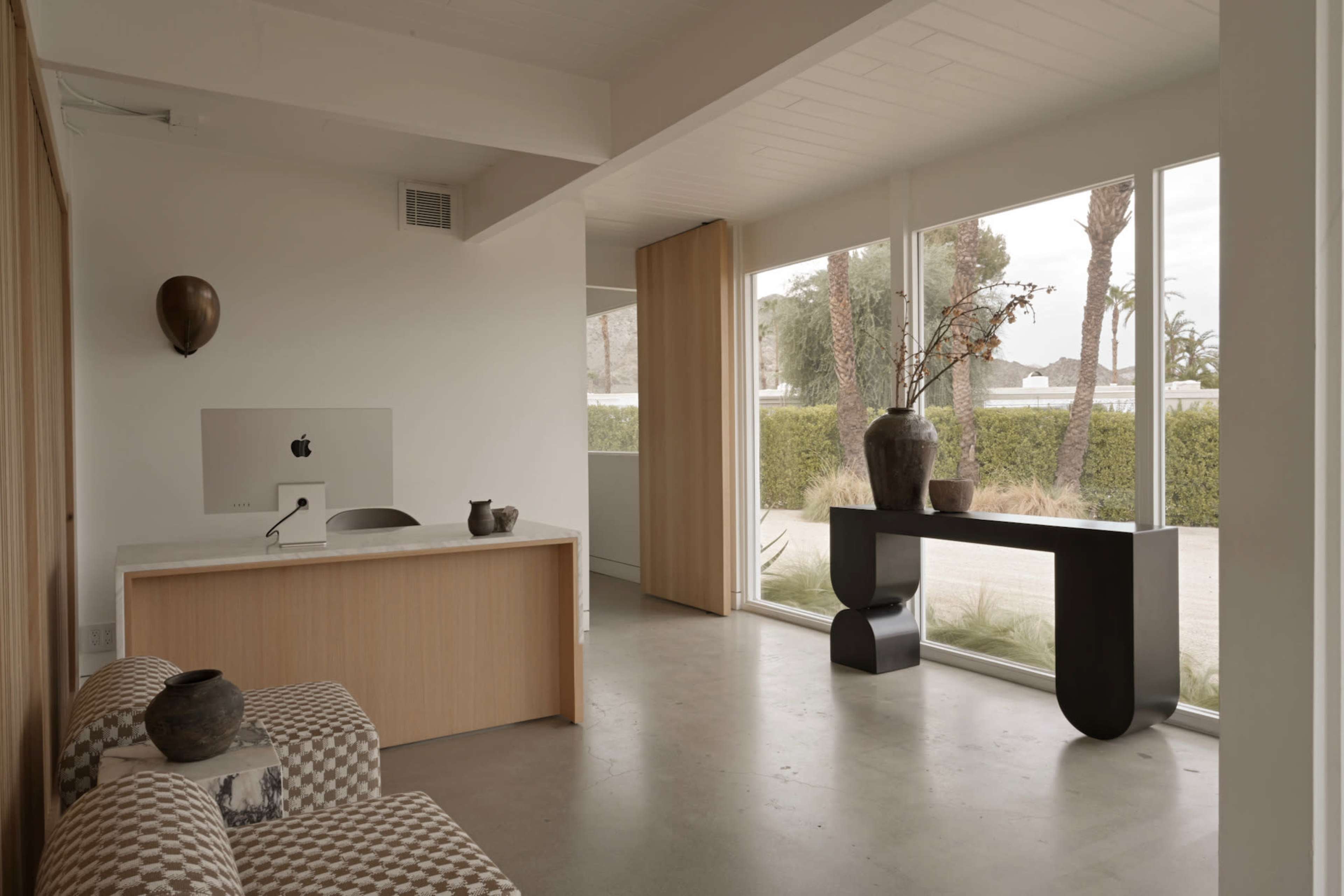 The image shows a modern office interior with a wooden reception desk, a computer, a minimalist black console table, and large windows that allow natural light to fill the space.