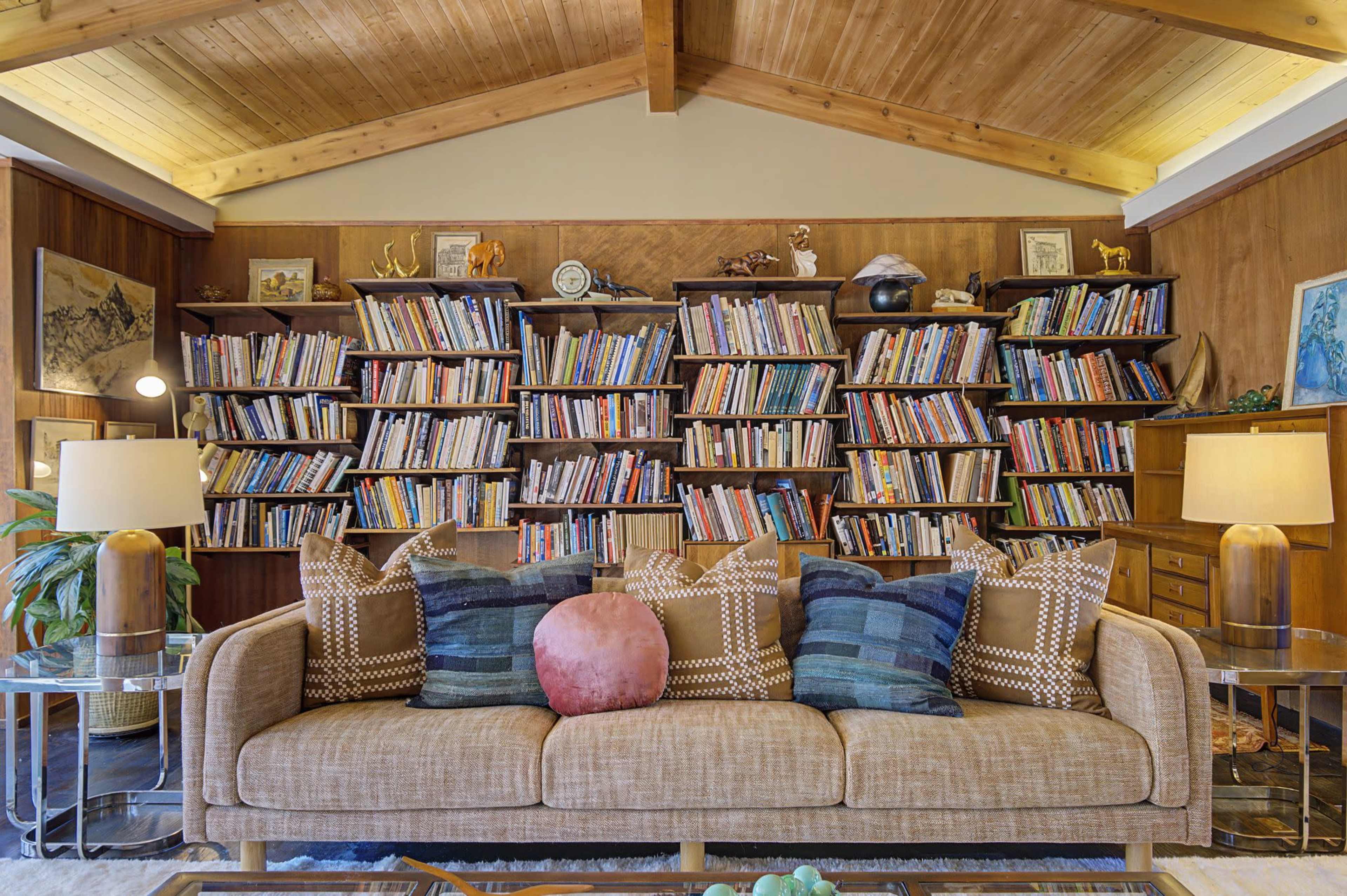 The image shows a living room with a beige sofa adorned with throw pillows, facing a large bookshelf filled with numerous books, under a wooden vaulted ceiling.