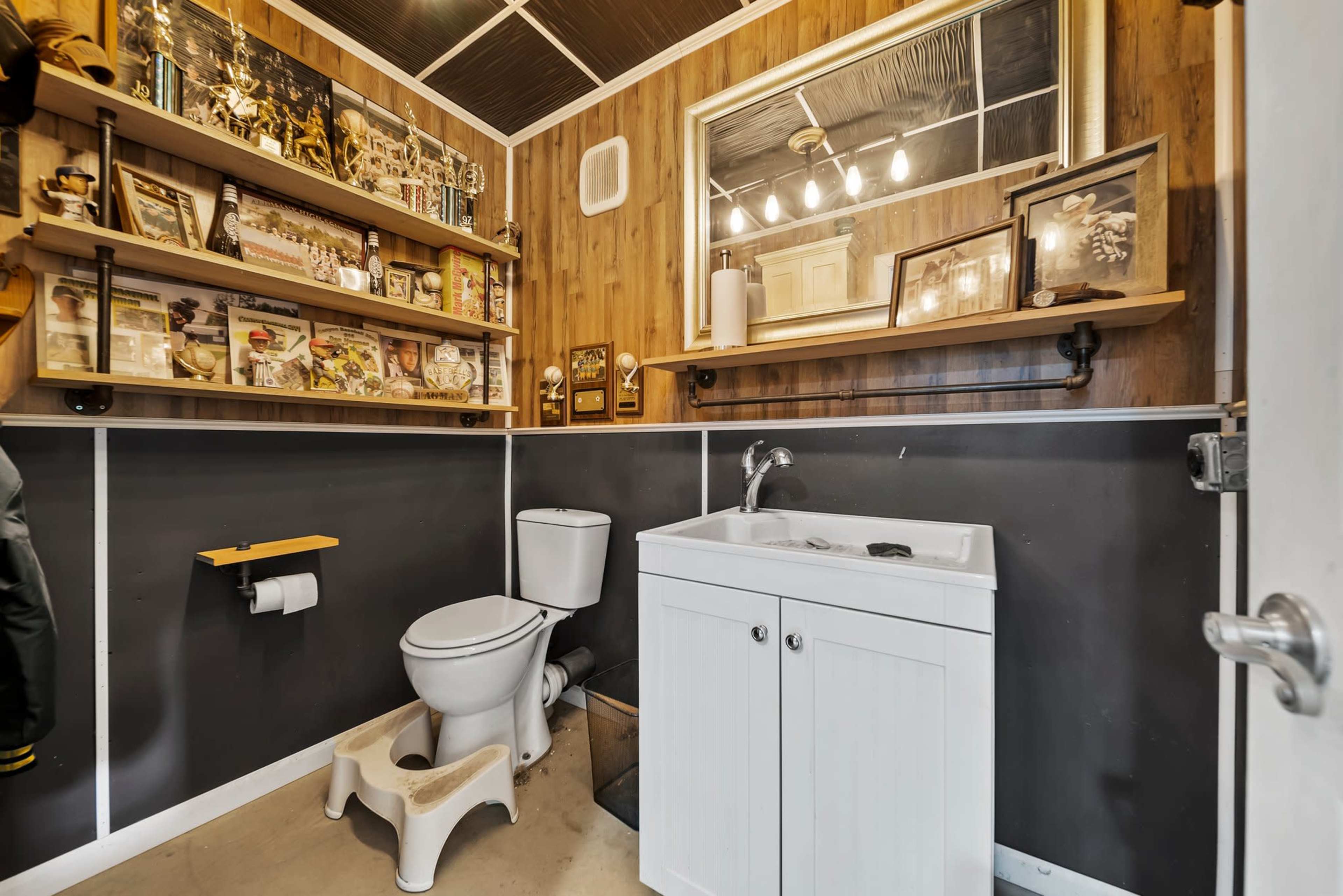 The image shows a bathroom featuring a toilet, a small sink with a cabinet, and shelves displaying various framed photos and memorabilia on the walls.