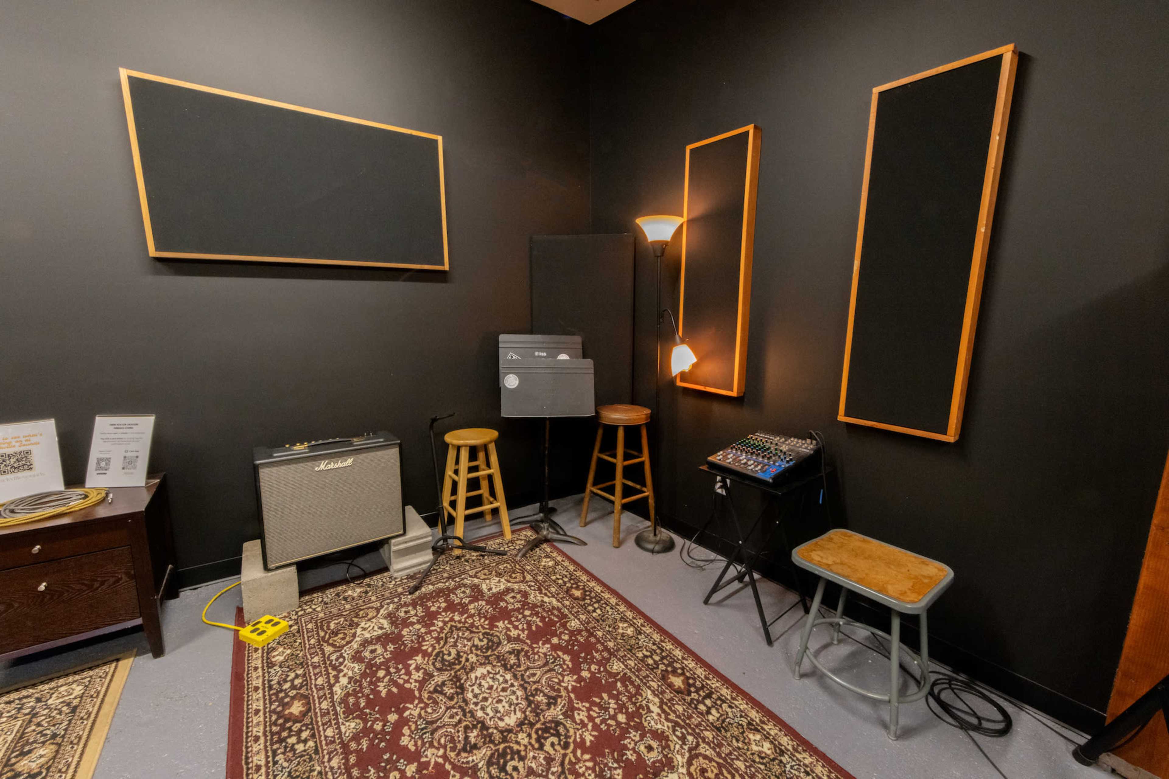 A small music rehearsal room with acoustic panels on the walls, a vintage amplifier, two stools, and a mixing console on a metal stool, all set on a patterned rug.