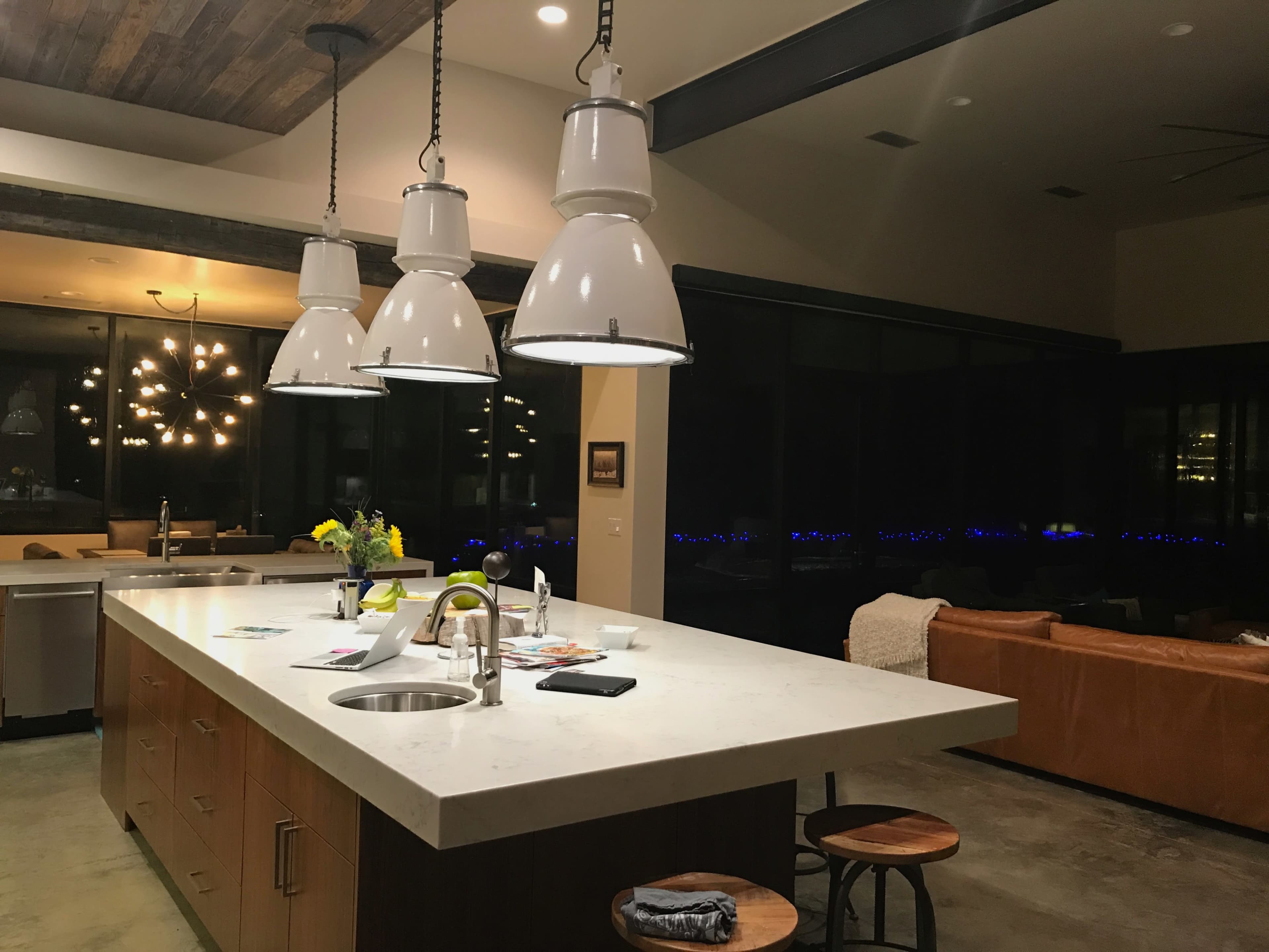 The image shows a modern kitchen with a central island, featuring three white pendant lights, wooden cabinets, and a large window revealing a night view.