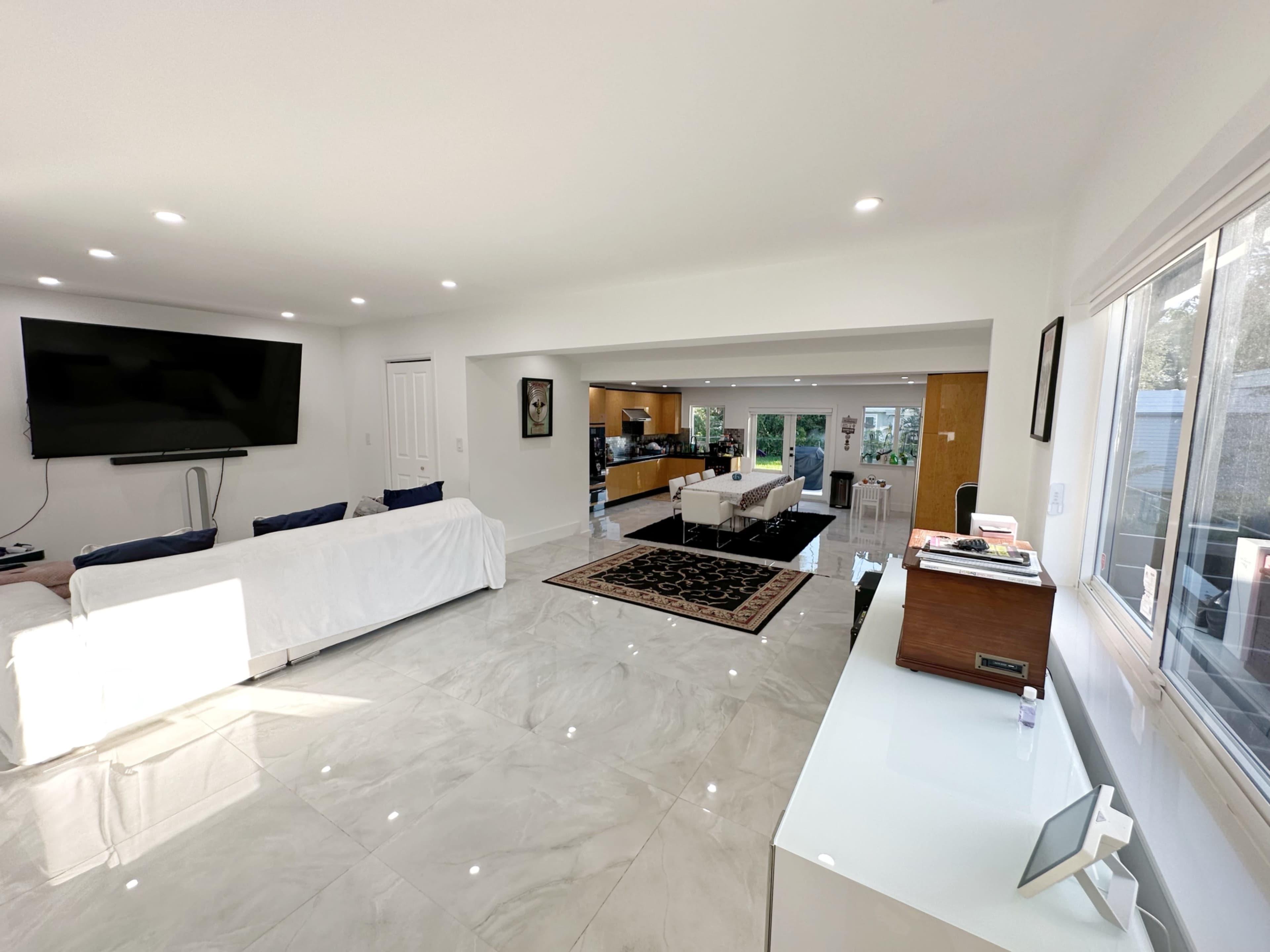A spacious, well-lit living room with a large television mounted on the wall, white furniture, and marble flooring leading to an open kitchen area.