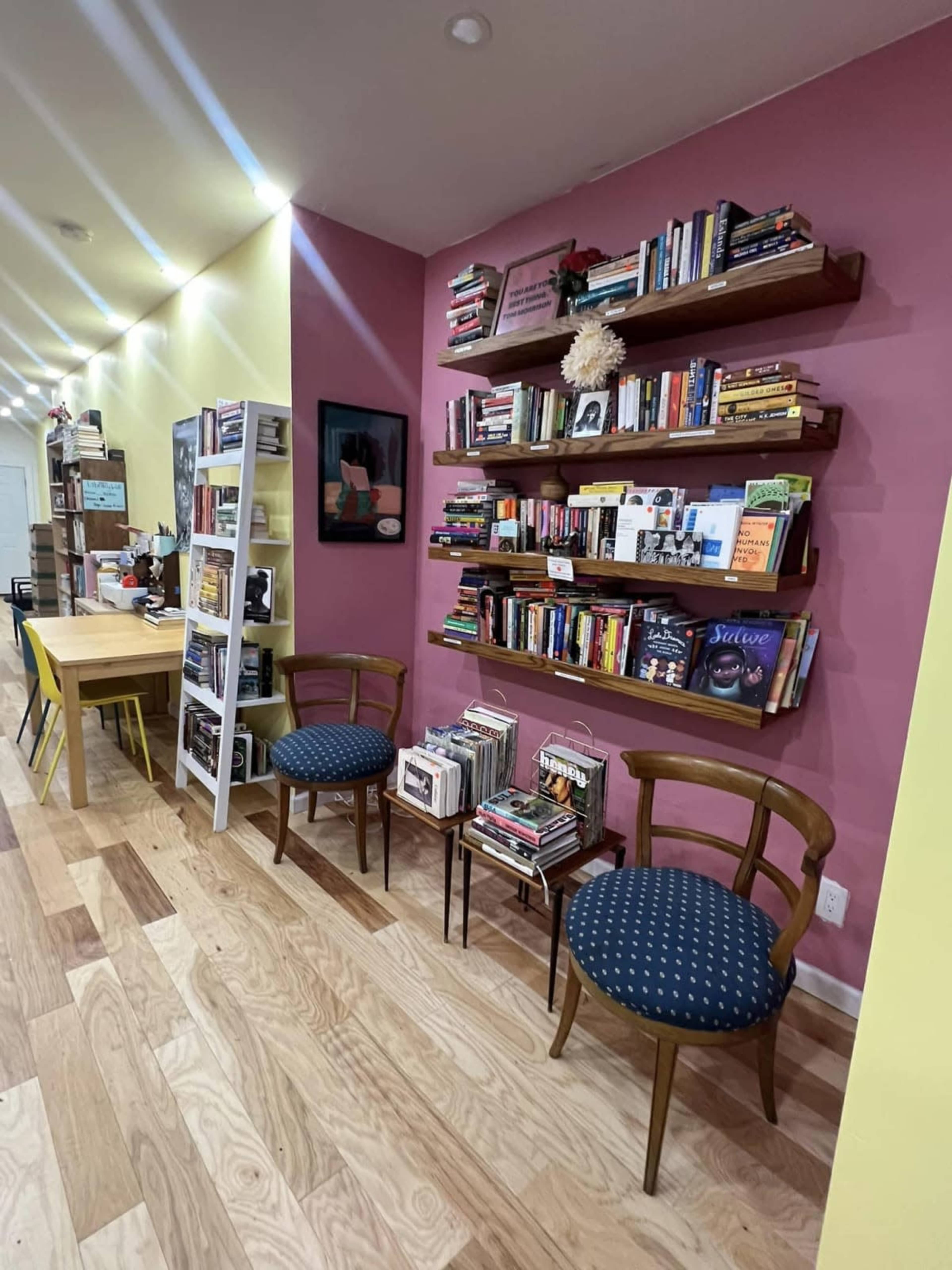 The image shows a cozy reading nook featuring two wooden chairs beside small tables stacked with books, against a pink wall adorned with floating bookshelves filled with various titles.