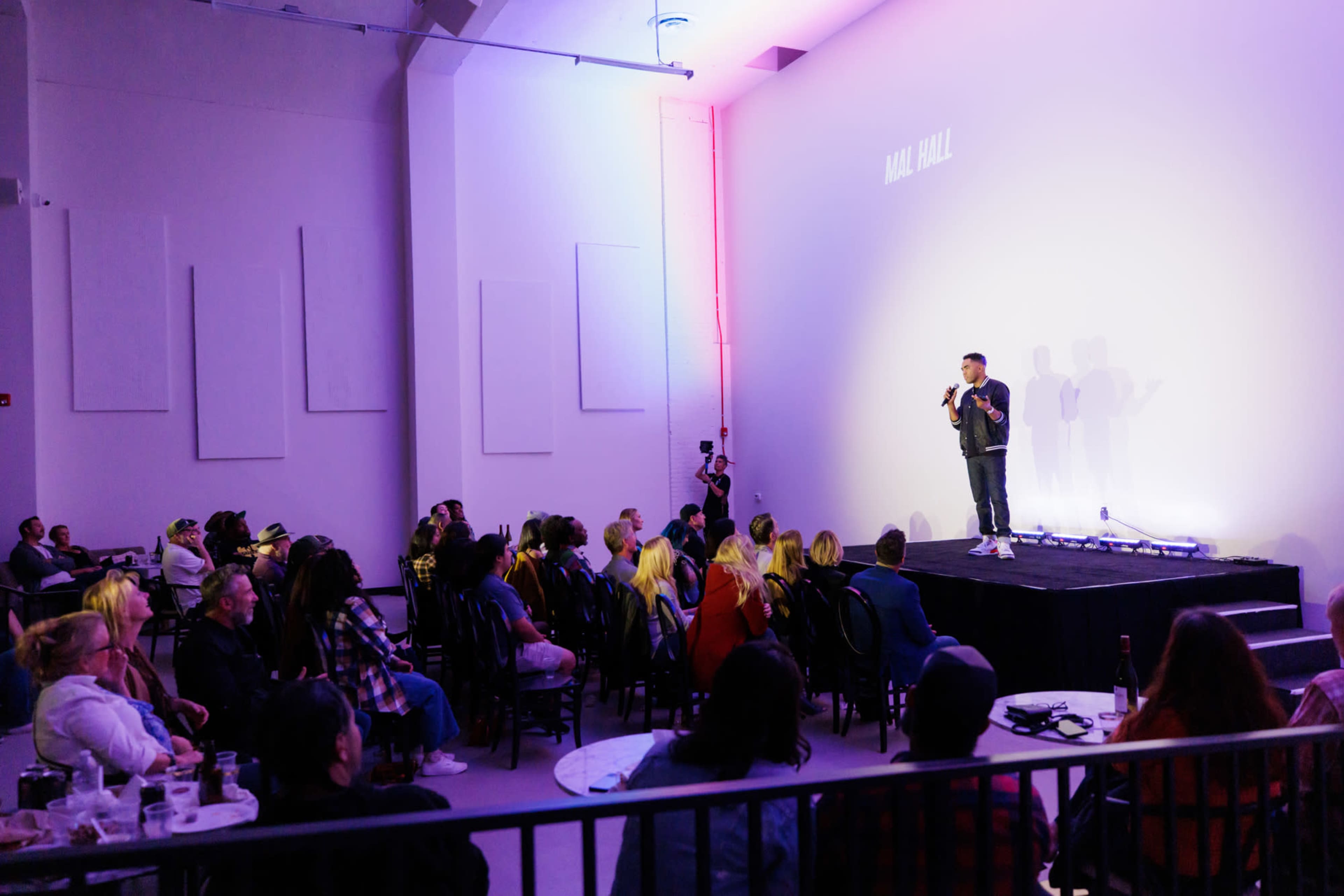 A speaker stands on stage in a modern venue while an audience observes from seated tables.