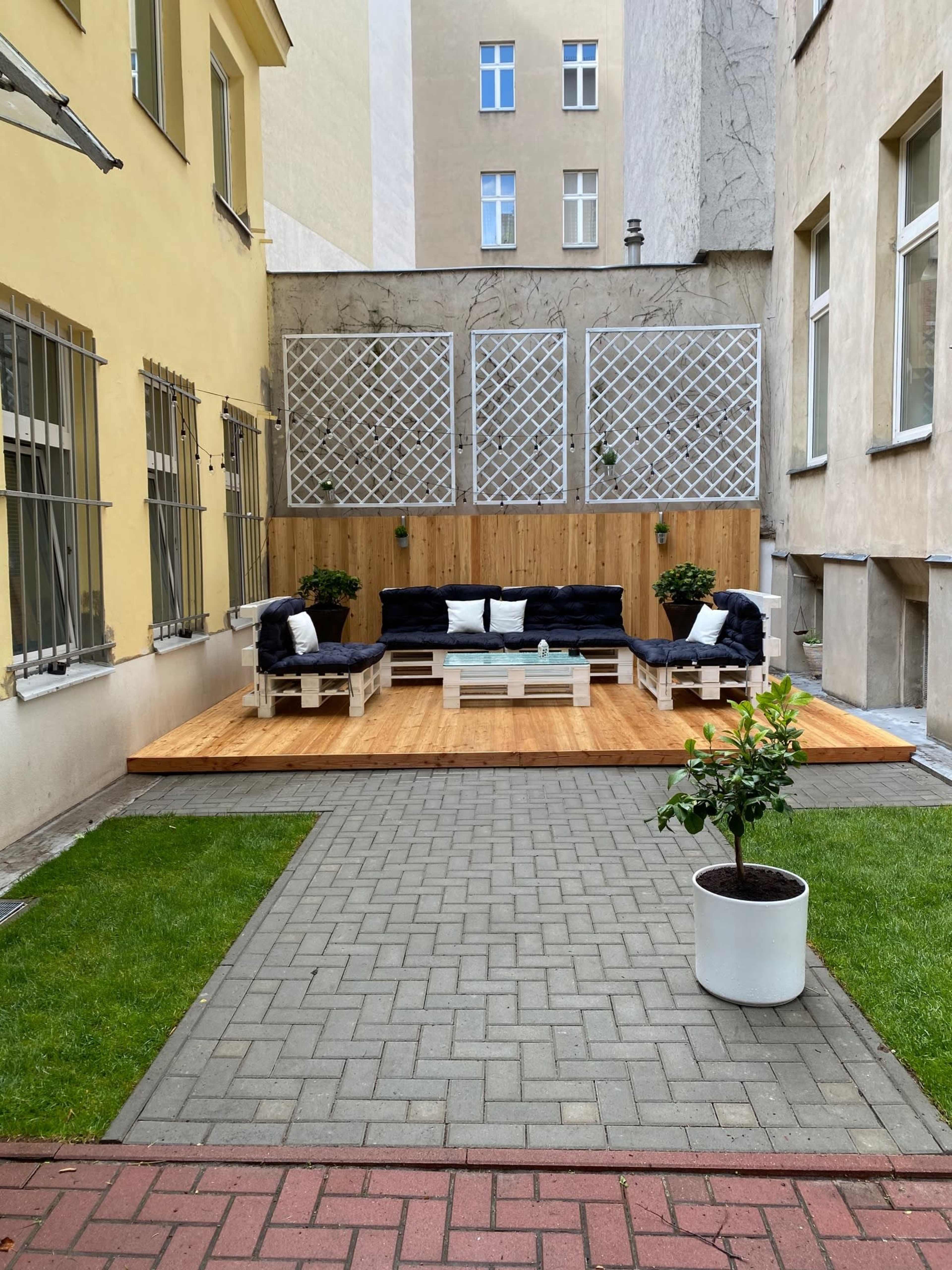 A modern outdoor seating area with wooden decking, black couches, a central table, and plants, enclosed by two beige building walls.