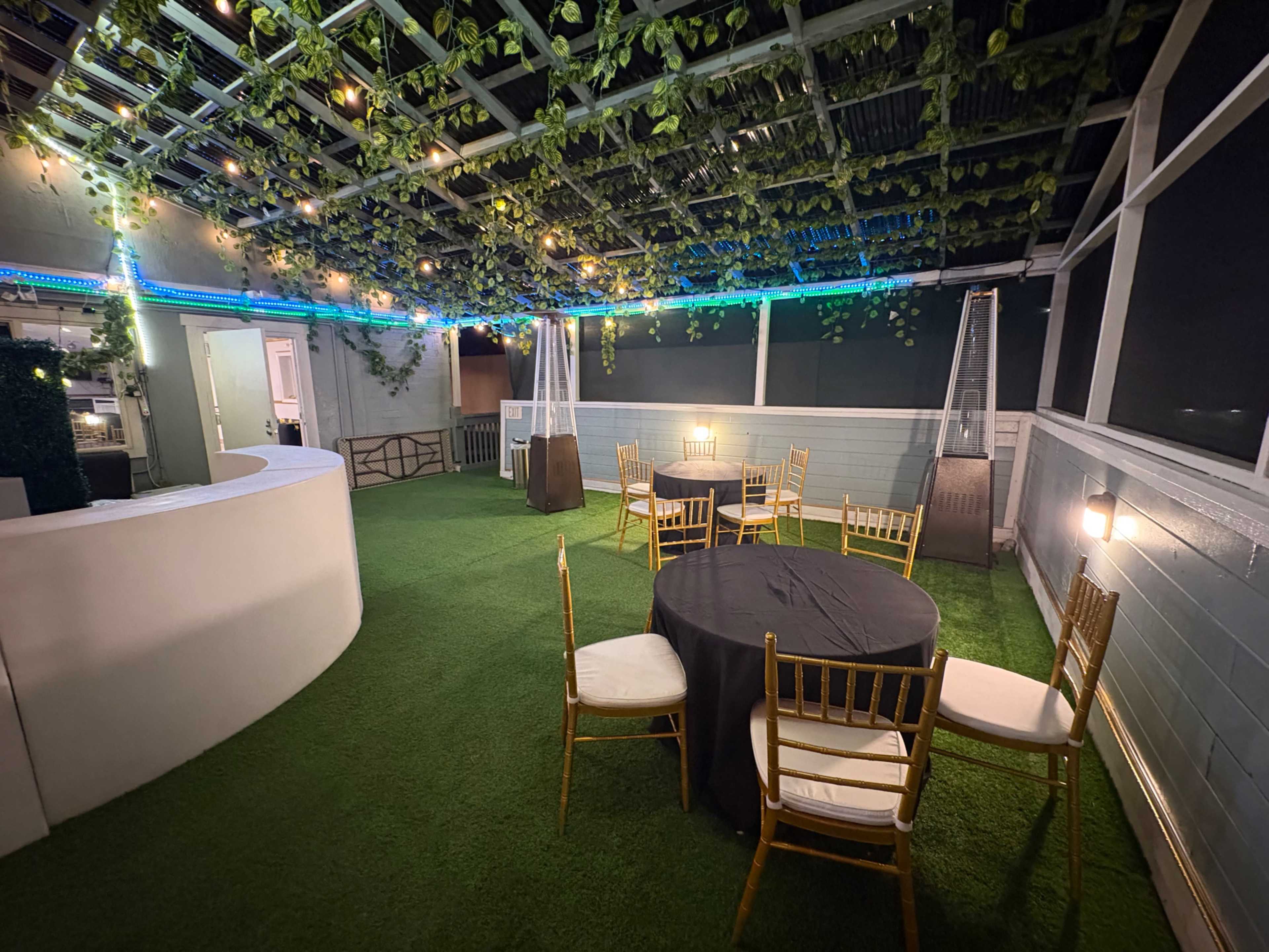 The image shows a rooftop event space with green artificial grass, round tables covered in black cloth, and hanging plants illuminated by soft lights.