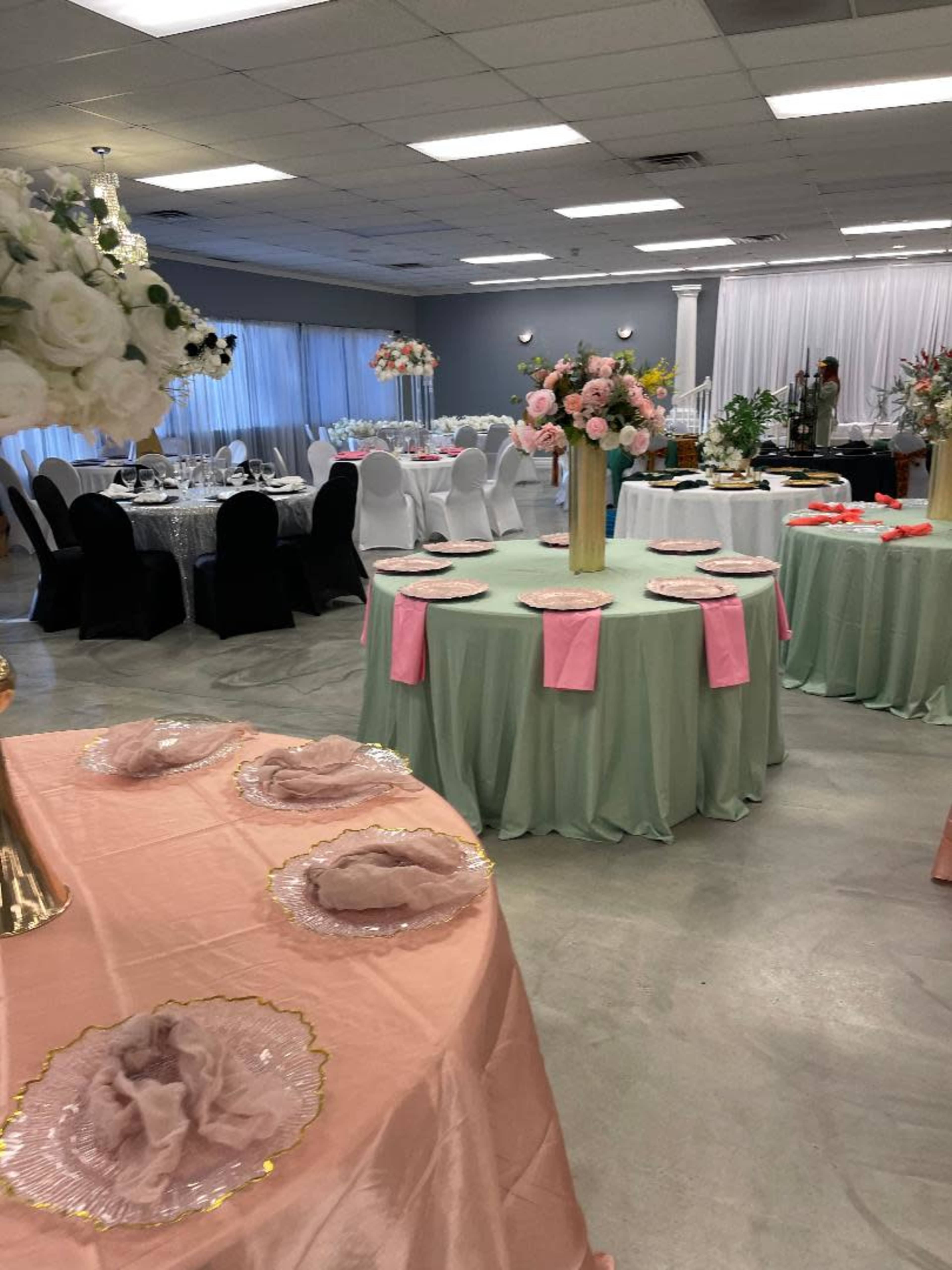 The image shows a banquet hall set up for an event, featuring elegantly decorated tables with pastel-colored tablecloths, floral centerpieces, and a mixture of round and rectangular seating arrangements.