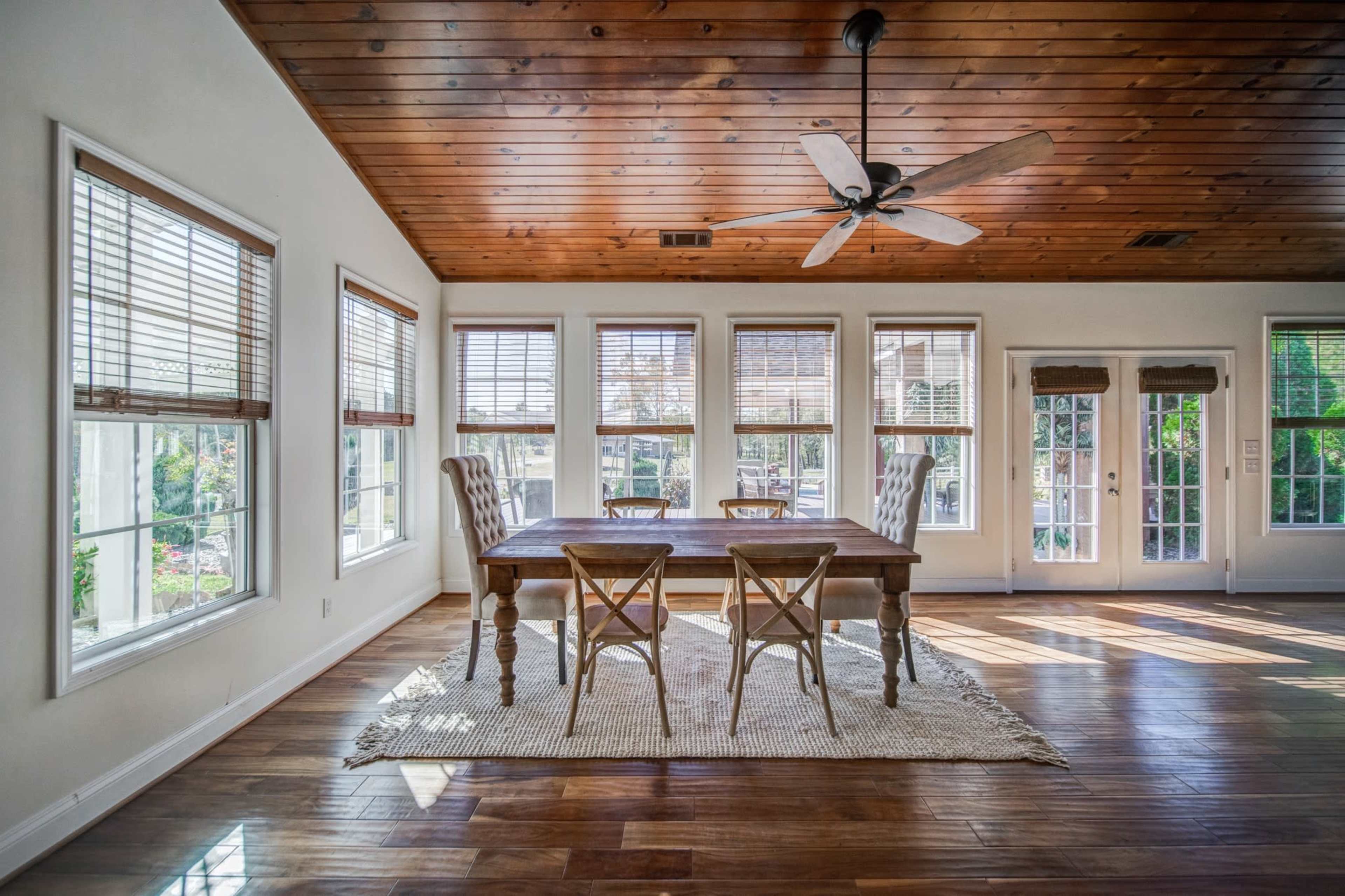 A wooden dining table with four chairs is positioned in a bright room featuring large windows, a ceiling fan, and a hardwood floor.