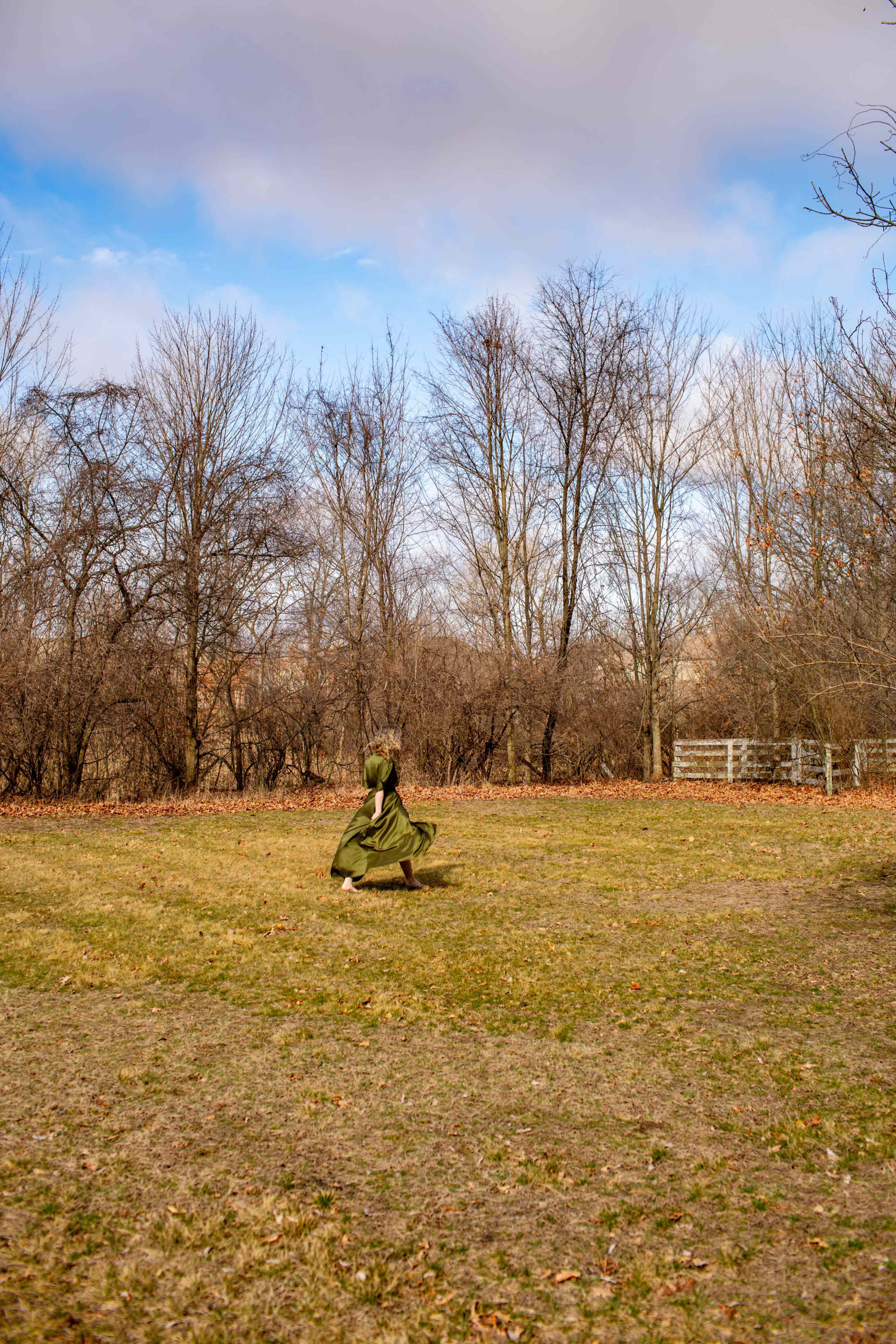 A person wearing a green garment is running across a grassy field surrounded by bare trees under a partly cloudy sky.