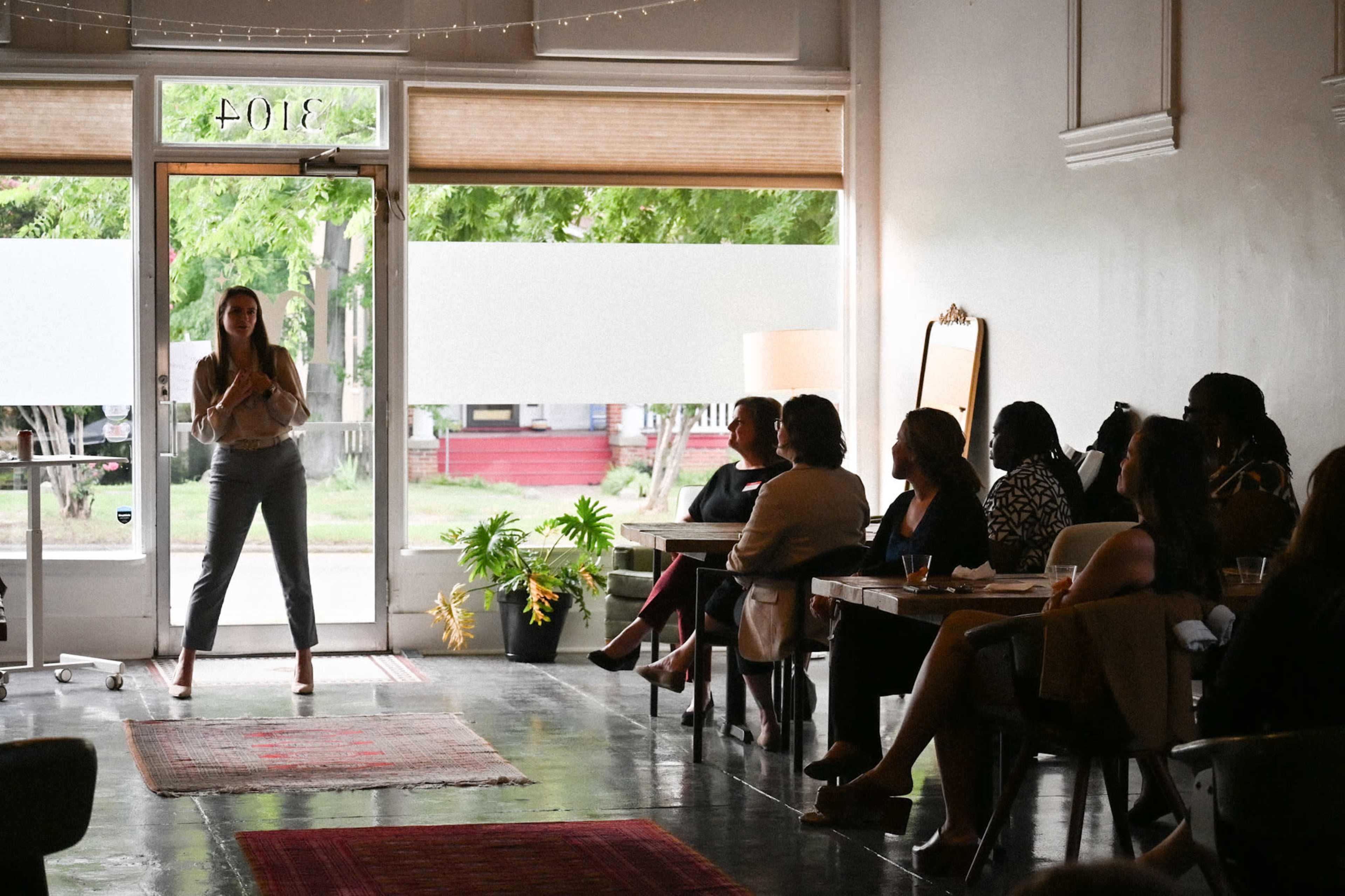 A woman stands in front of a group seated at tables in a light-filled room, while the audience attentively listens.