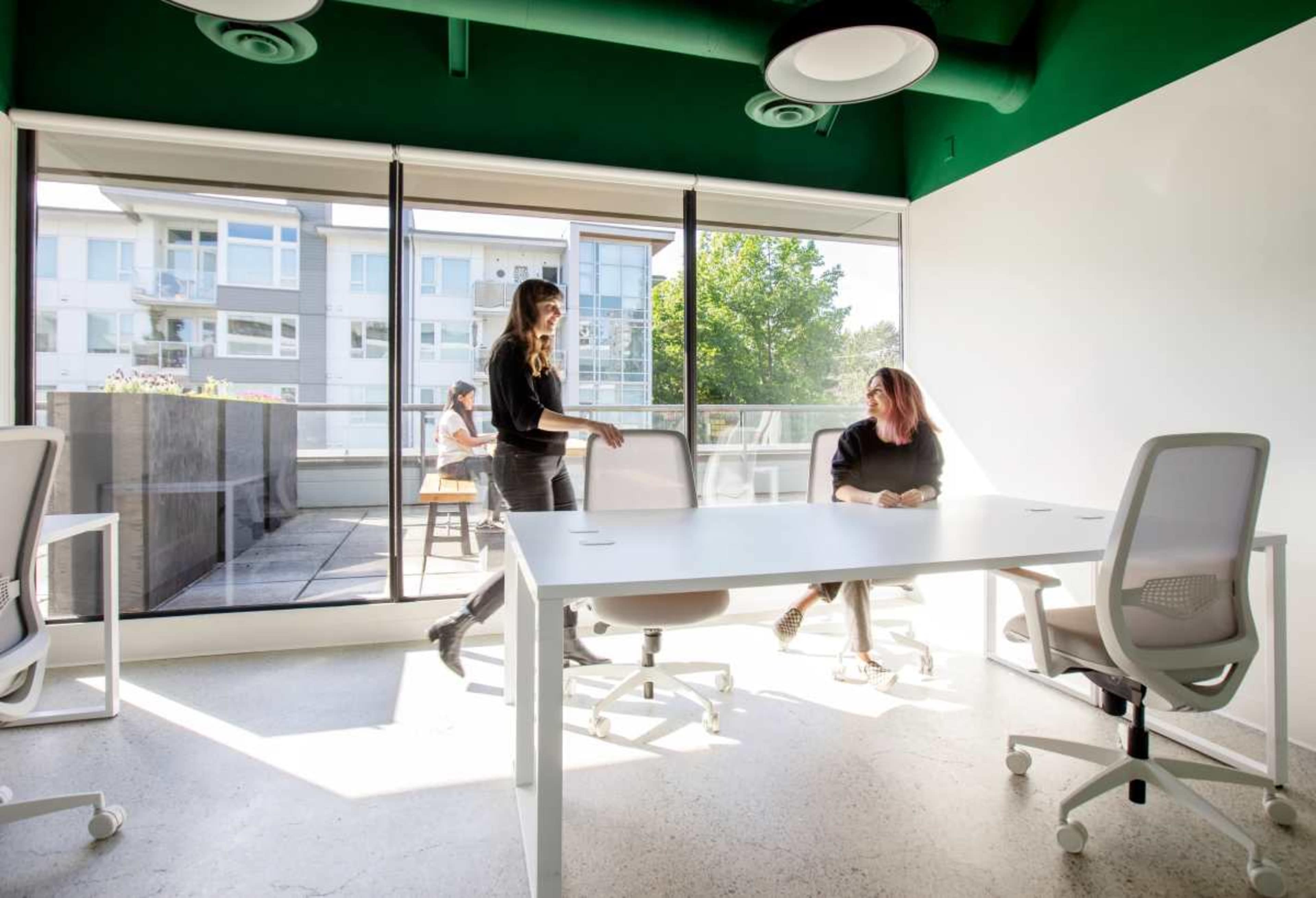 Two women are engaged in conversation in a bright office space with large windows and modern furniture.