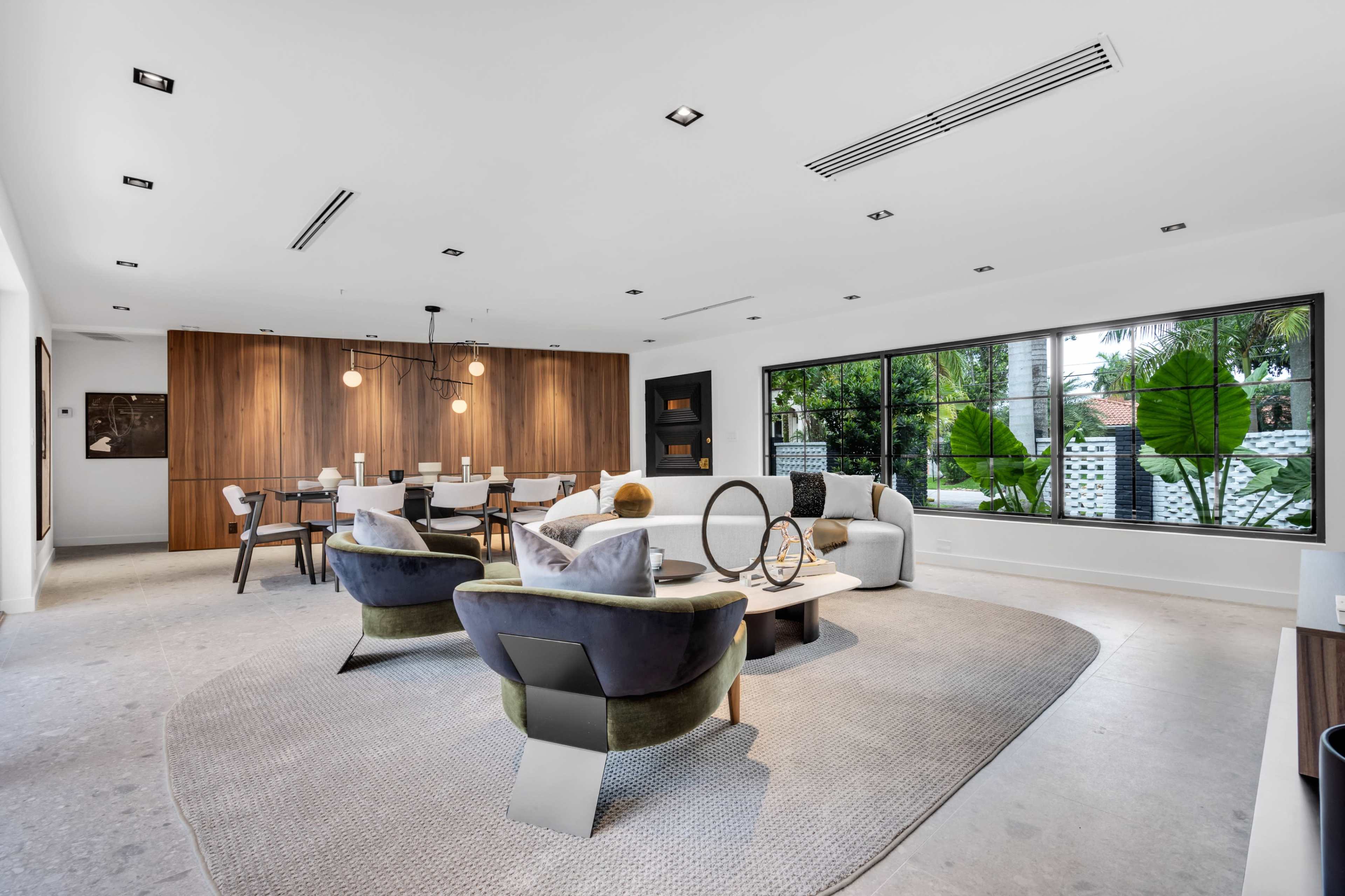A modern living room features a seating area with chairs and a coffee table, alongside a dining space with a wooden table and chairs, illuminated by pendant lighting.