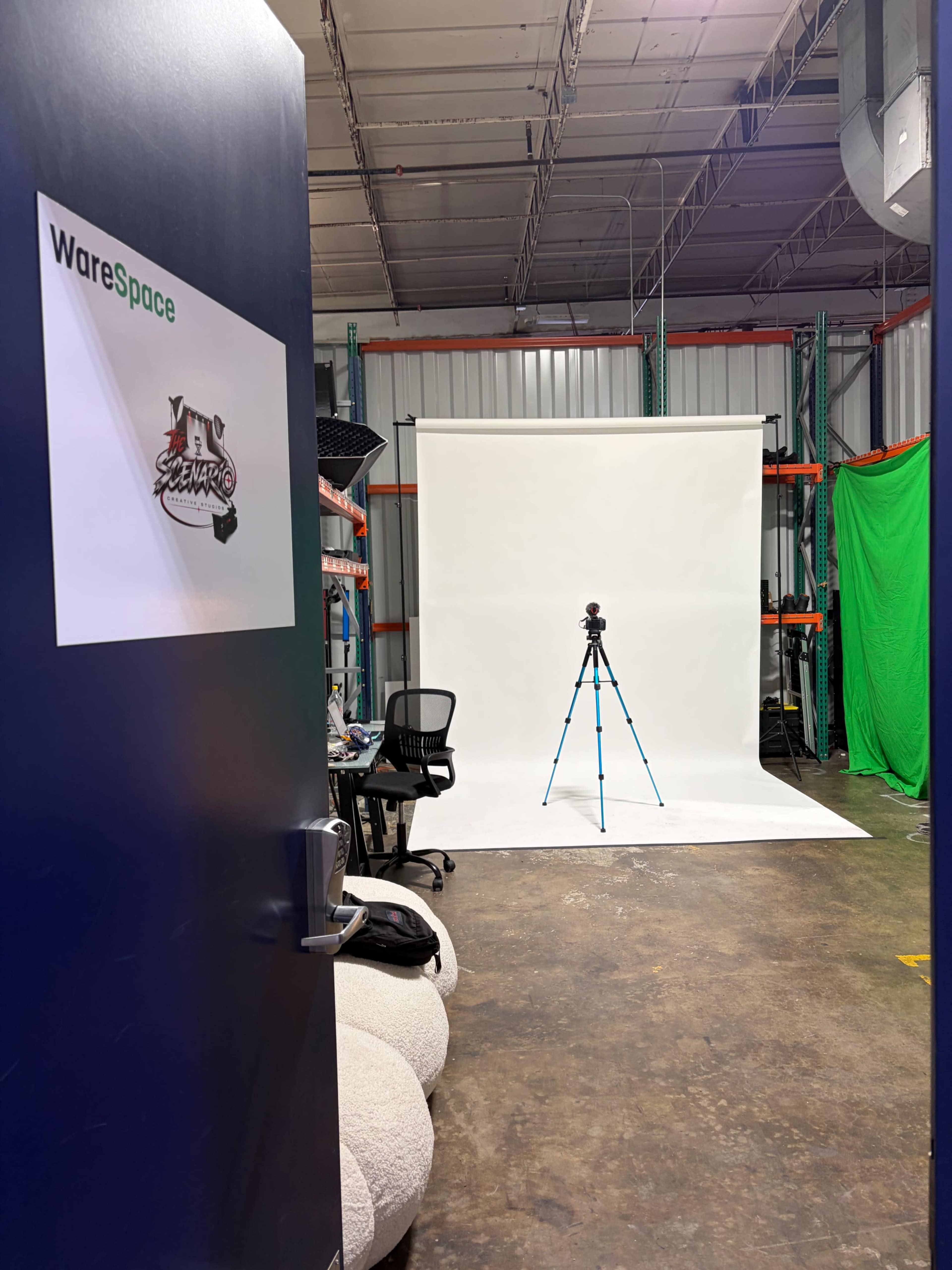 The image shows an open door leading to a space with a white backdrop, a tripod in the center, and green screen walls in a storage area.