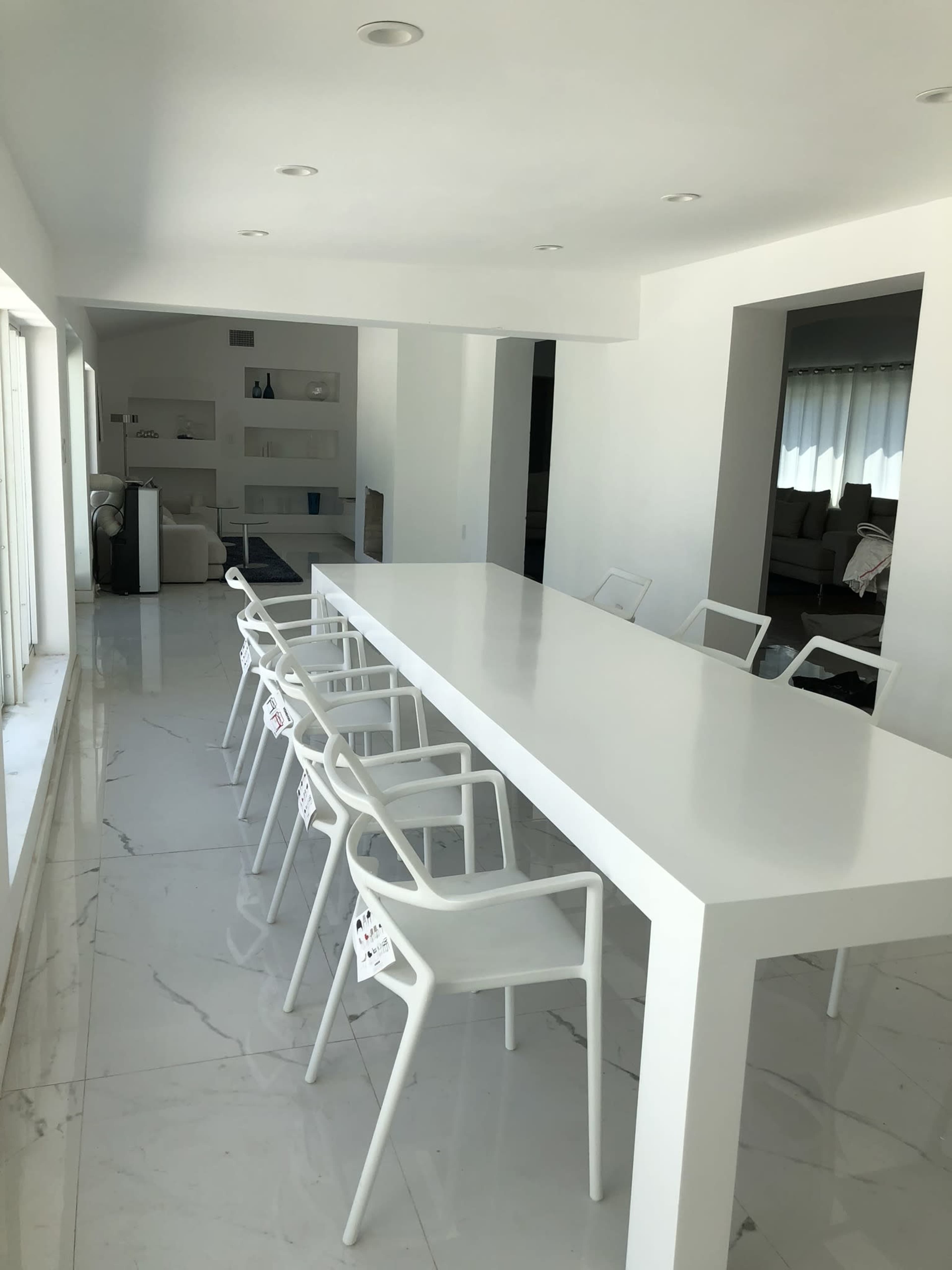 A modern dining area with a long white table and several white chairs, set against a backdrop of spacious white walls and marble flooring.