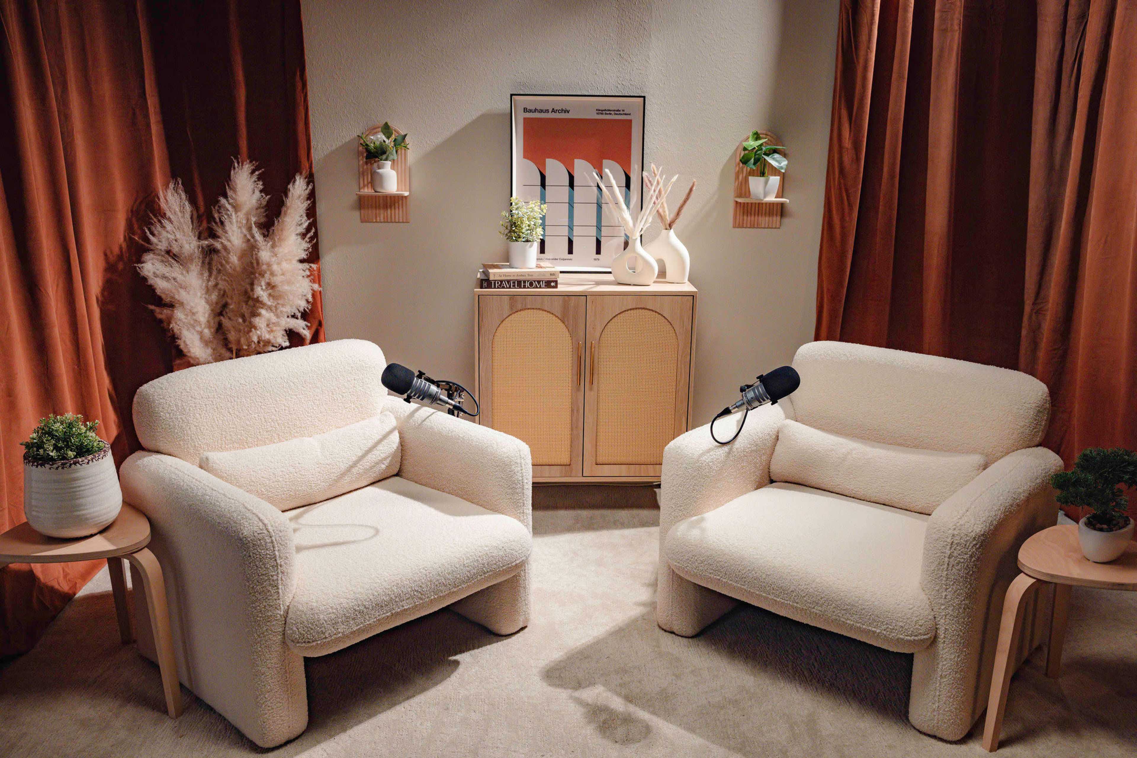 A cozy interior setting with two tufted armchairs, a wooden cabinet in the center, and microphones on each chair, surrounded by plants and decorative elements.
