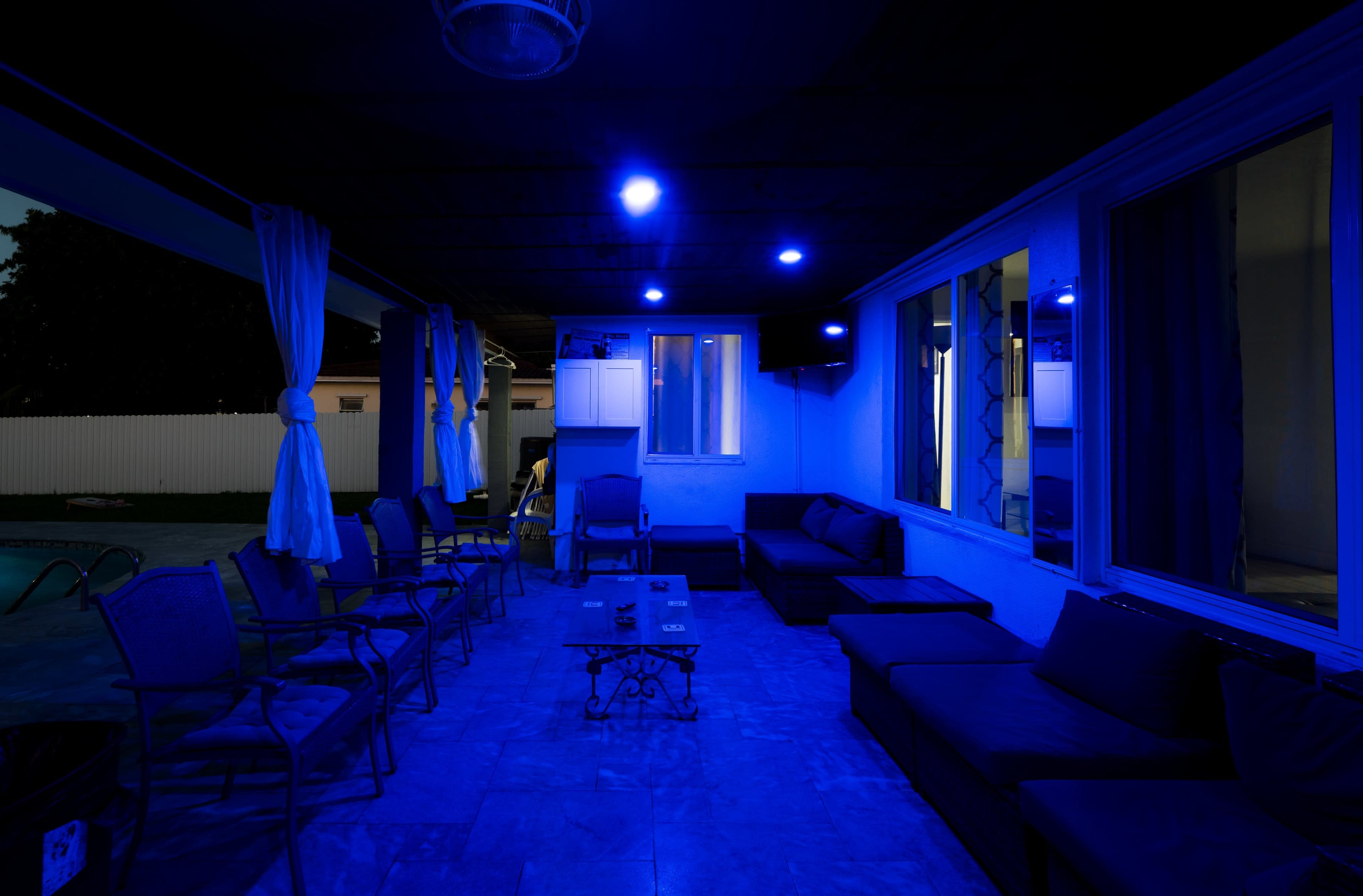 The image shows a dimly lit outdoor patio area with blue LED lights, featuring comfortable seating arrangements and a view of a swimming pool through an open space.