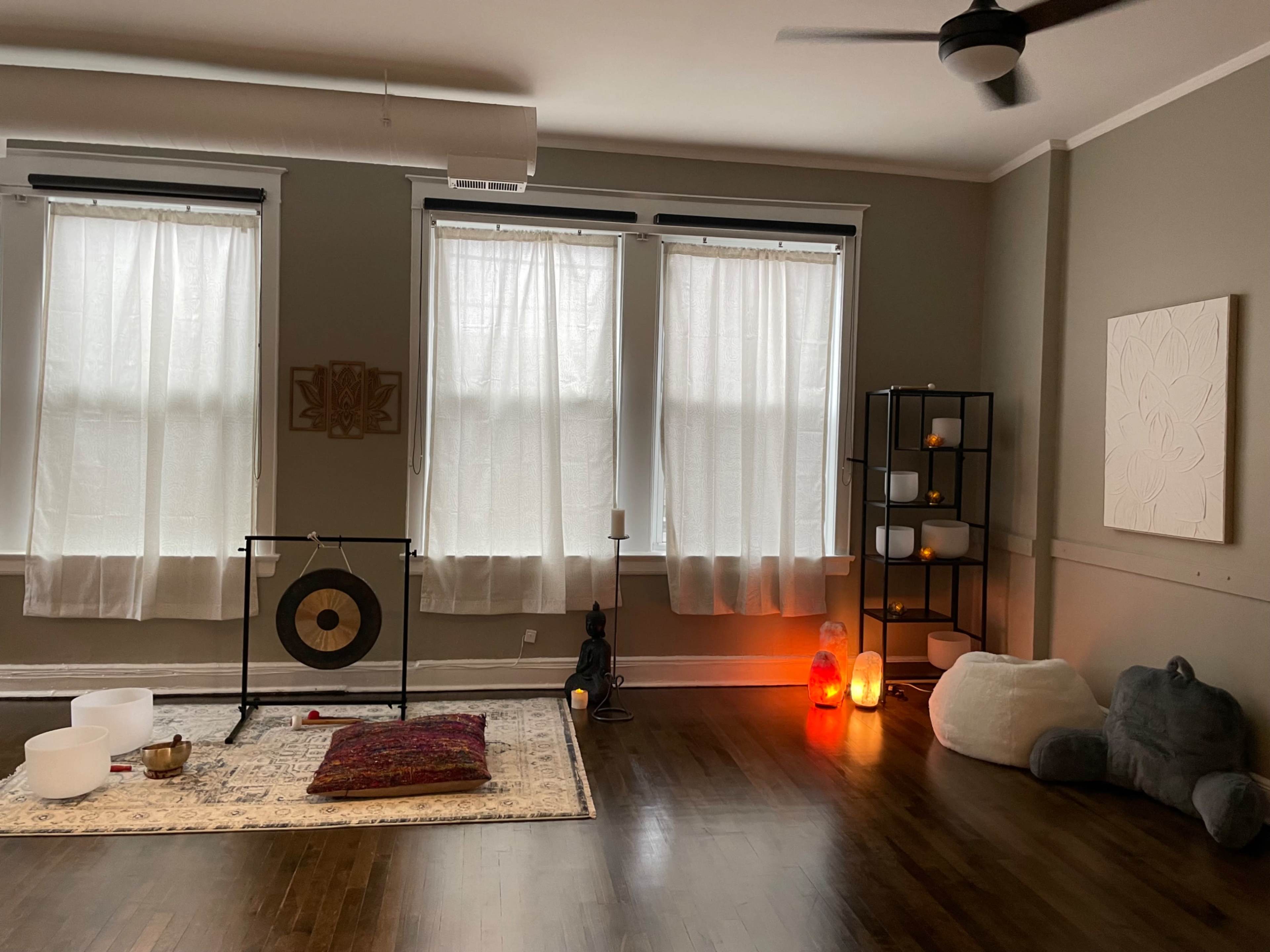 The room features three large windows with sheer curtains, a gong on a stand, various candles and lamps, a wall shelf with decorative items, and a soft seating area with floor cushions.