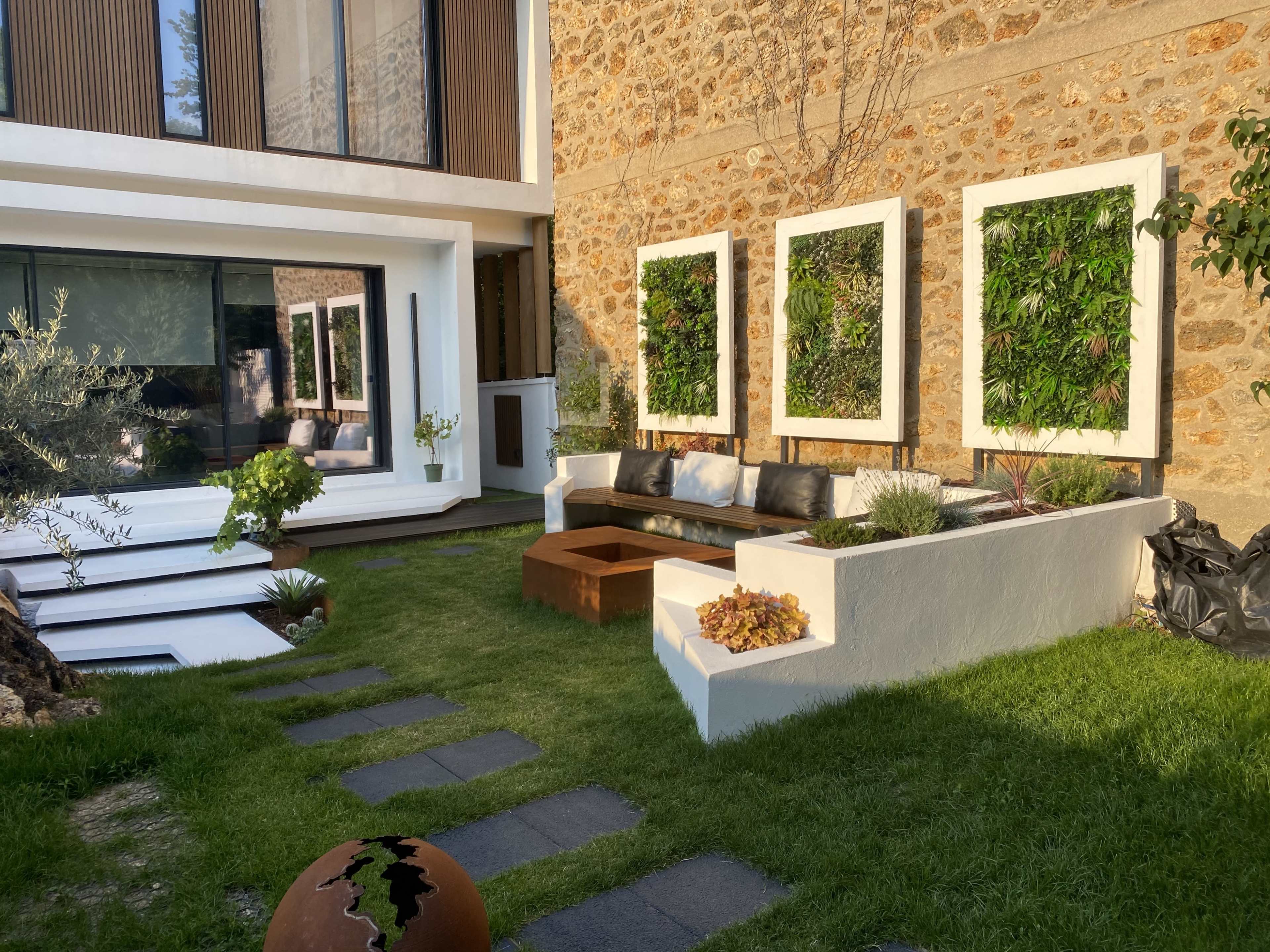 A modern outdoor living space with a seating area, planters, and framed vertical gardens, adjacent to a stone-walled house.