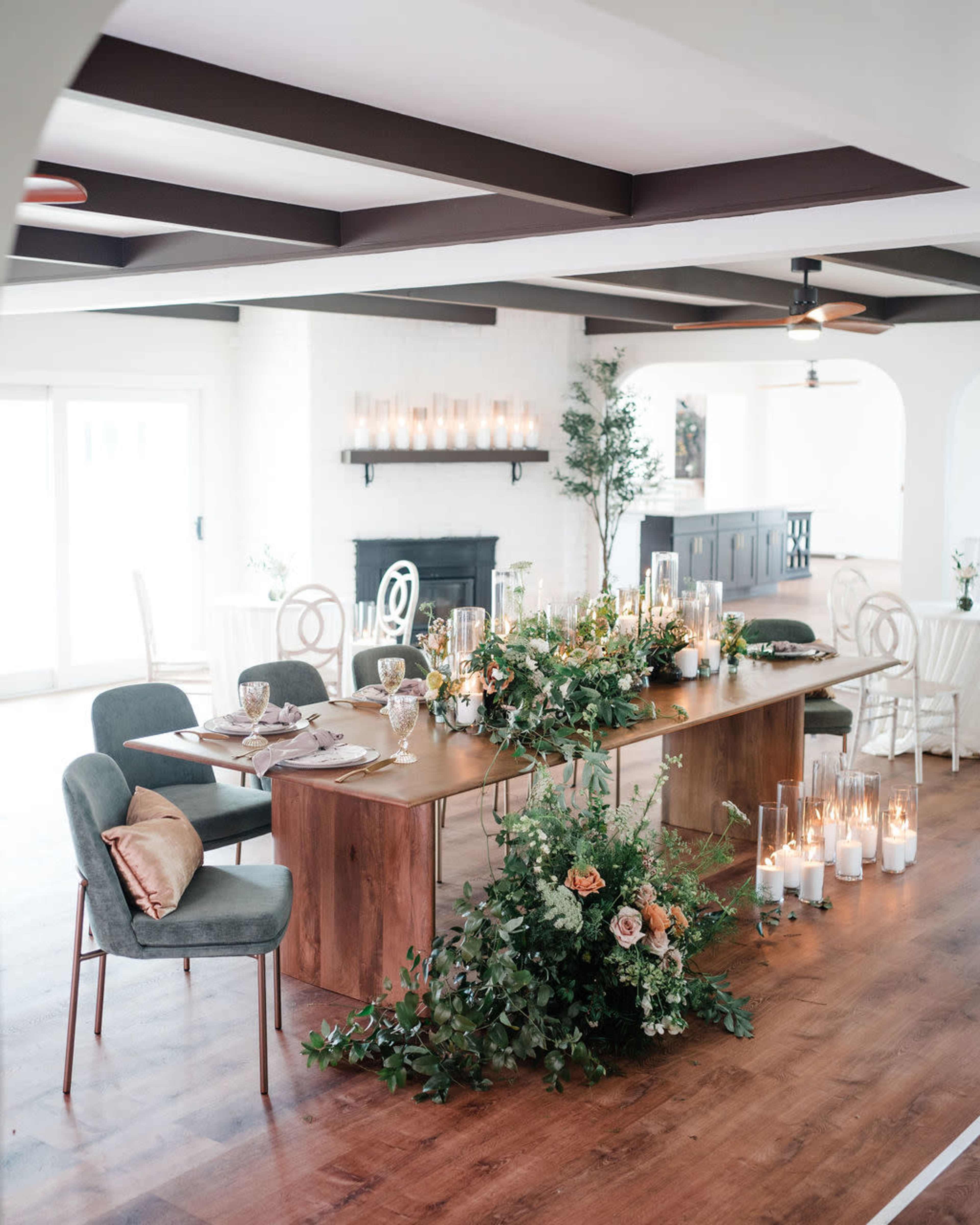 A long wooden table is set for a dining gathering, adorned with greenery and candles, in a bright, open room with modern decor.