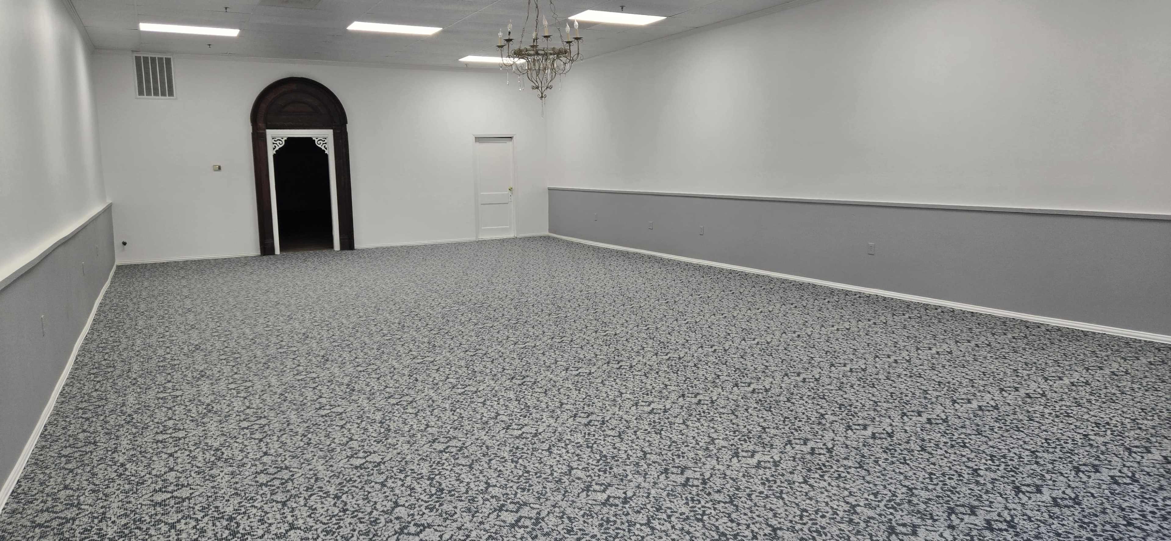 The image shows a large, empty room with patterned carpet, a chandelier hanging from the ceiling, and a door at one end leading to a darkened area.
