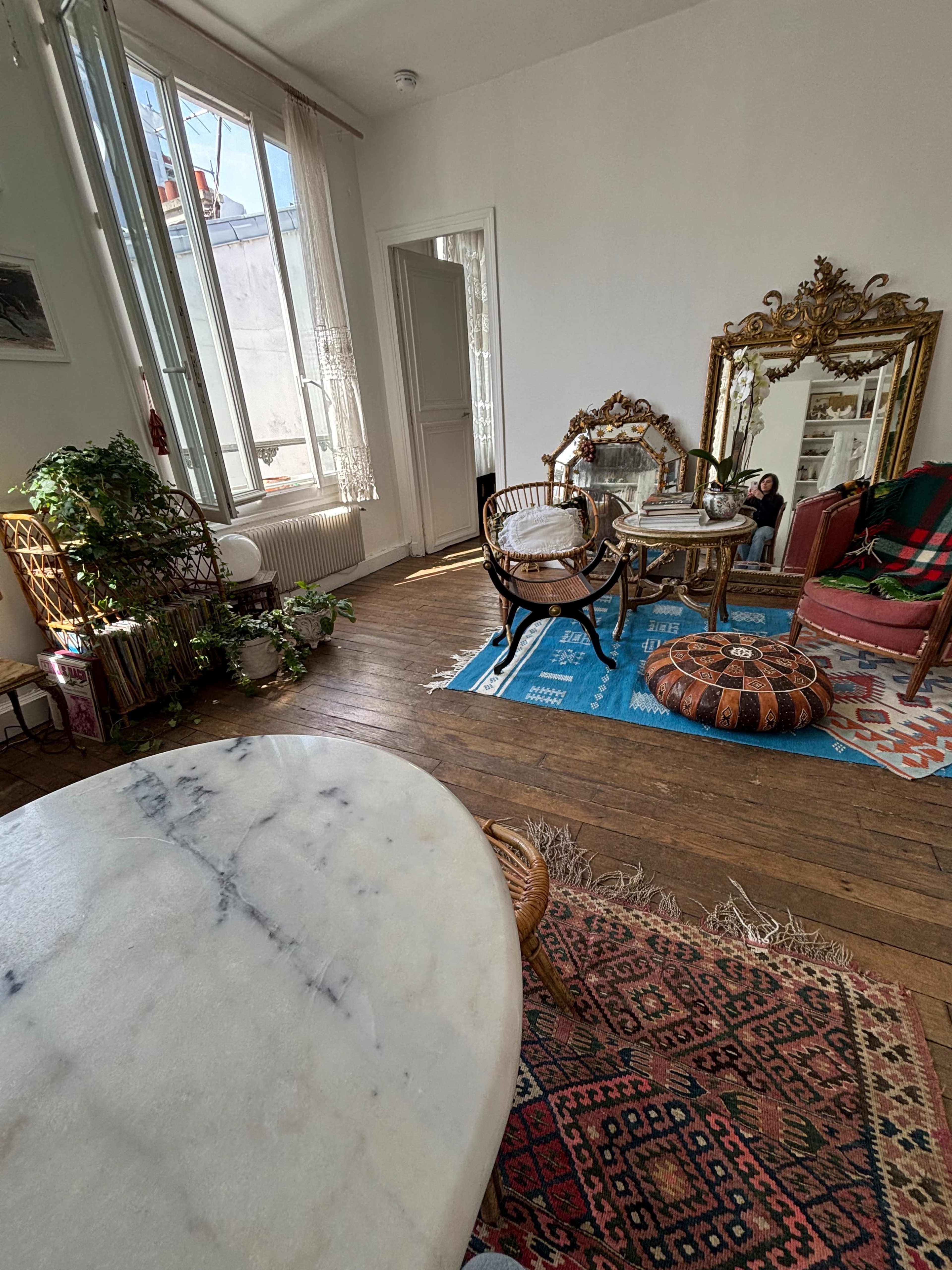 The image shows a sunlit room featuring a mix of vintage furniture, including a mirror, chairs, and a patterned rug, against a backdrop of white walls and large windows.