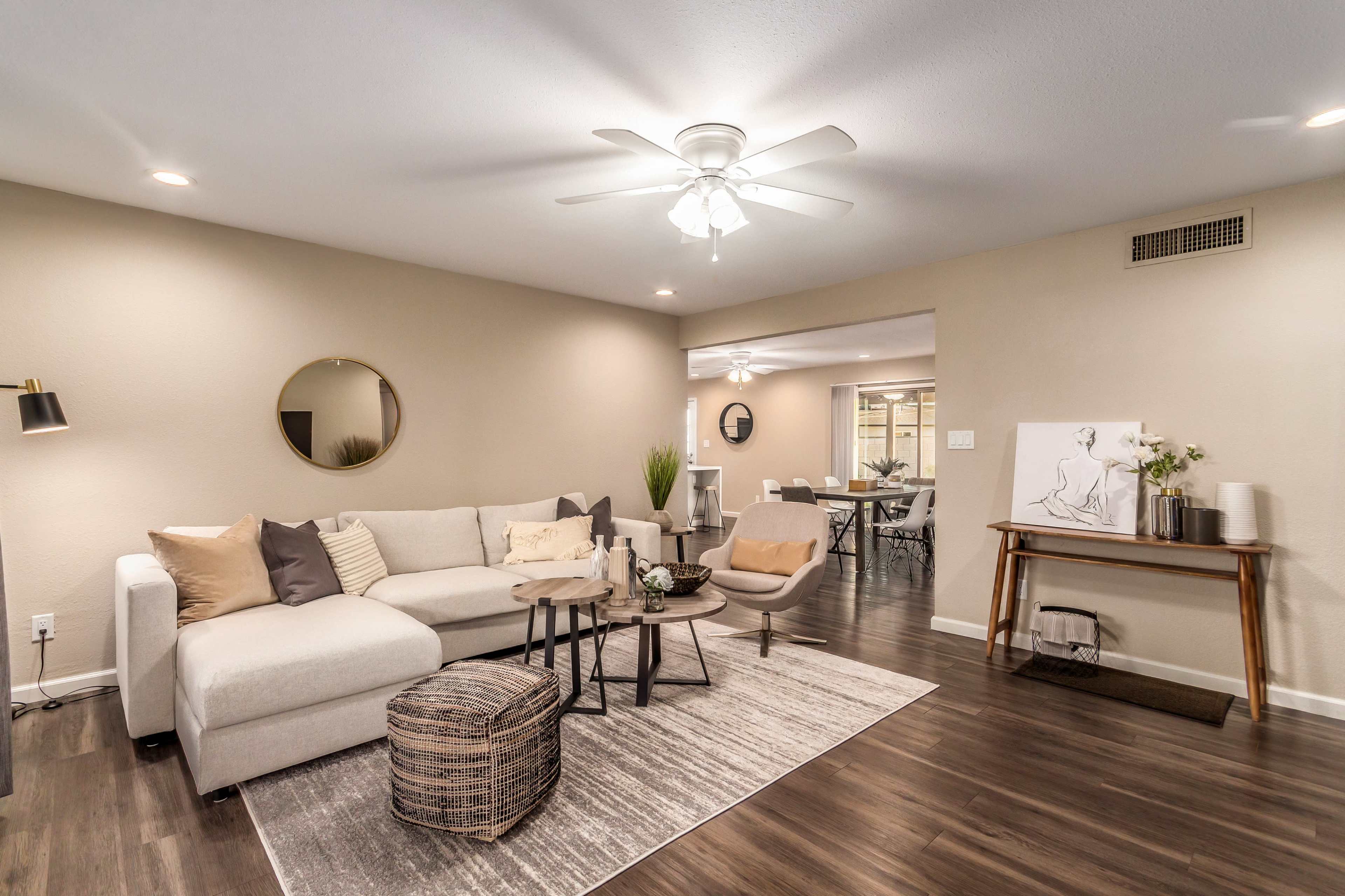 The image shows a modern living room featuring a light-colored sectional sofa, a round mirror on the wall, and a wooden side table with decorative items.