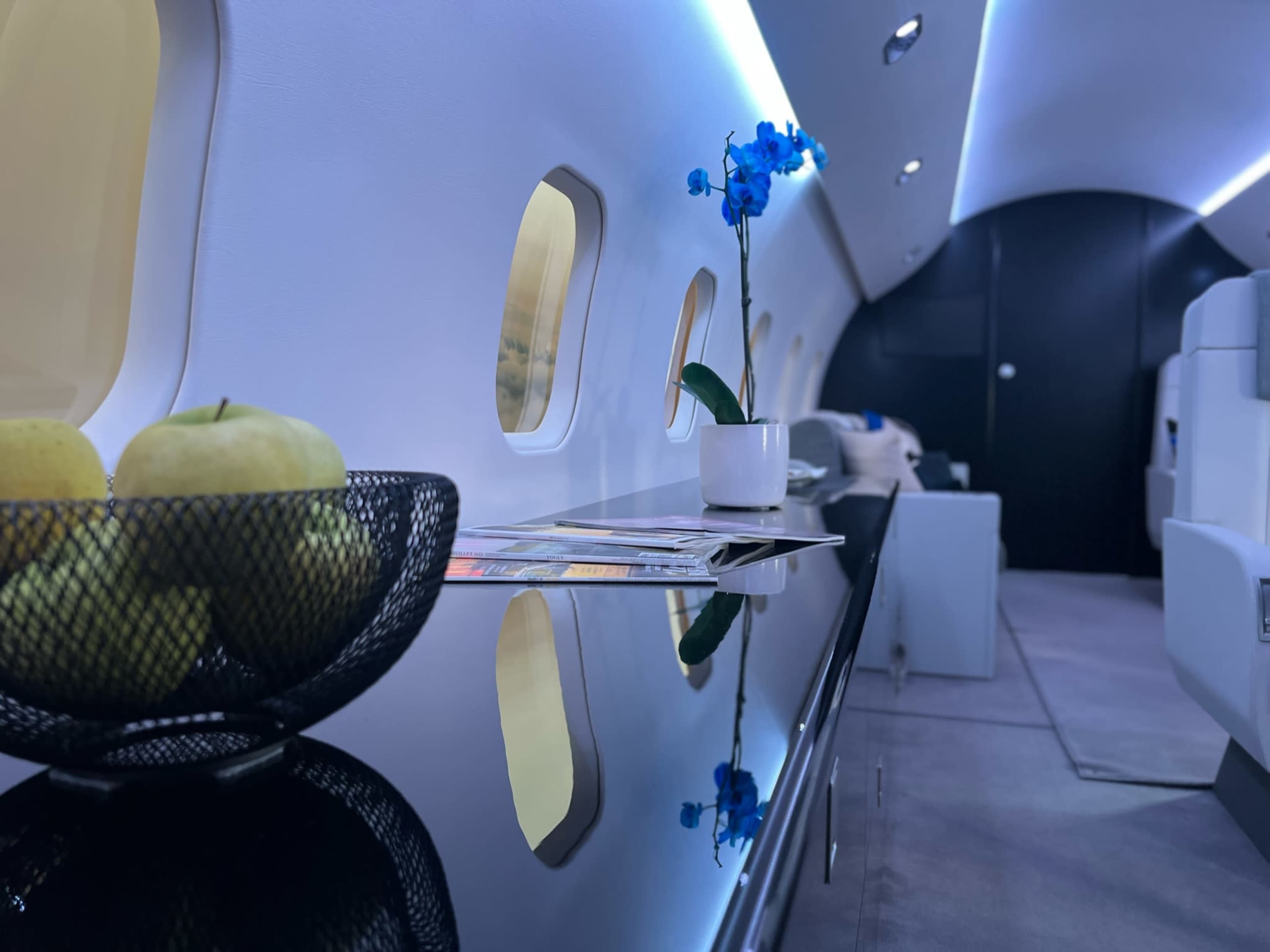 The interior of a private jet features a sleek black table with a bowl of green apples and a blue orchid in a pot, alongside illuminated windows and plush seating.