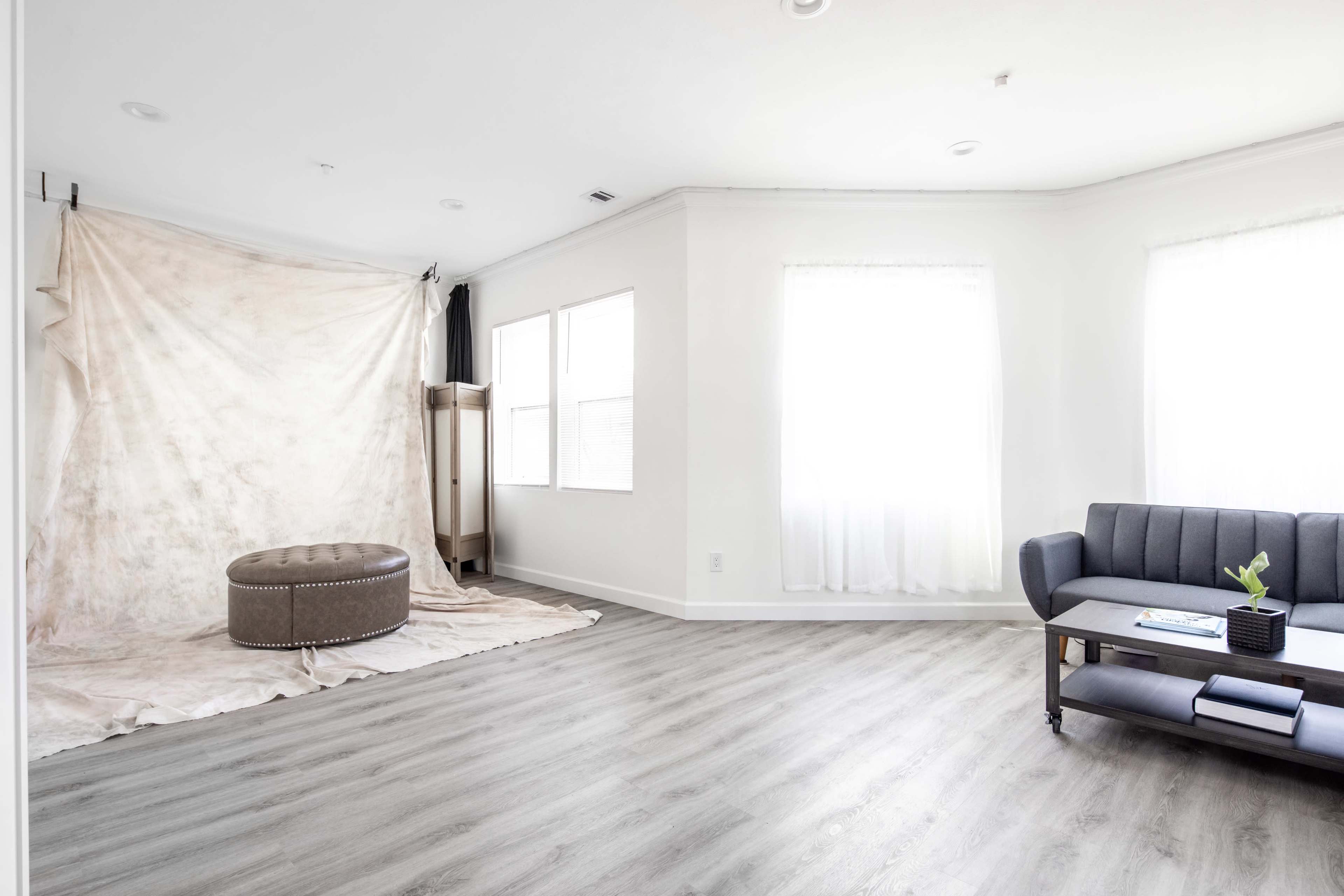 A bright, empty room with a light-colored backdrop, a round ottoman, and a gray sofa positioned near a window.