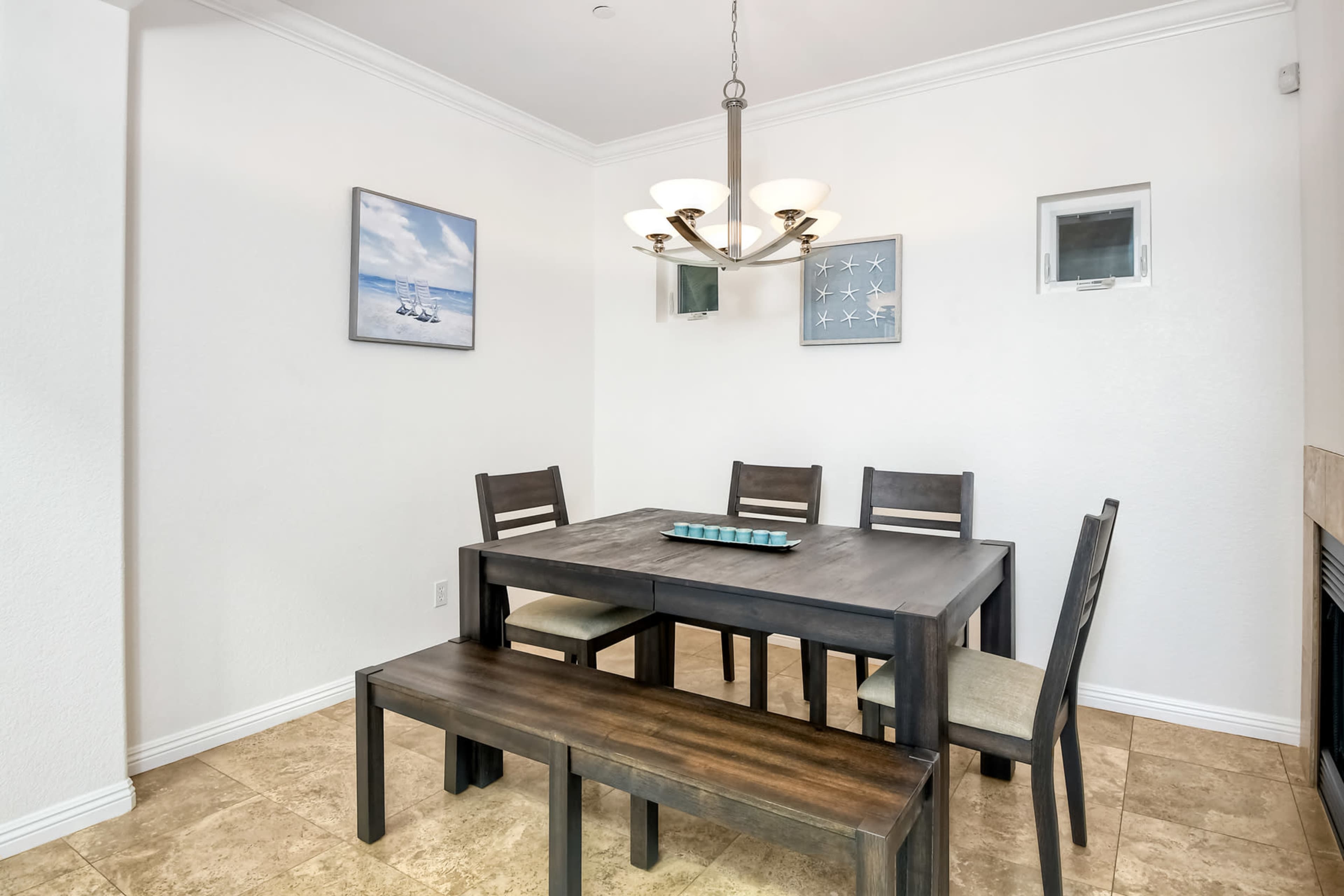 A dining area features a wooden table with six chairs and a bench, illuminated by a chandelier, set against white walls with framed artwork.