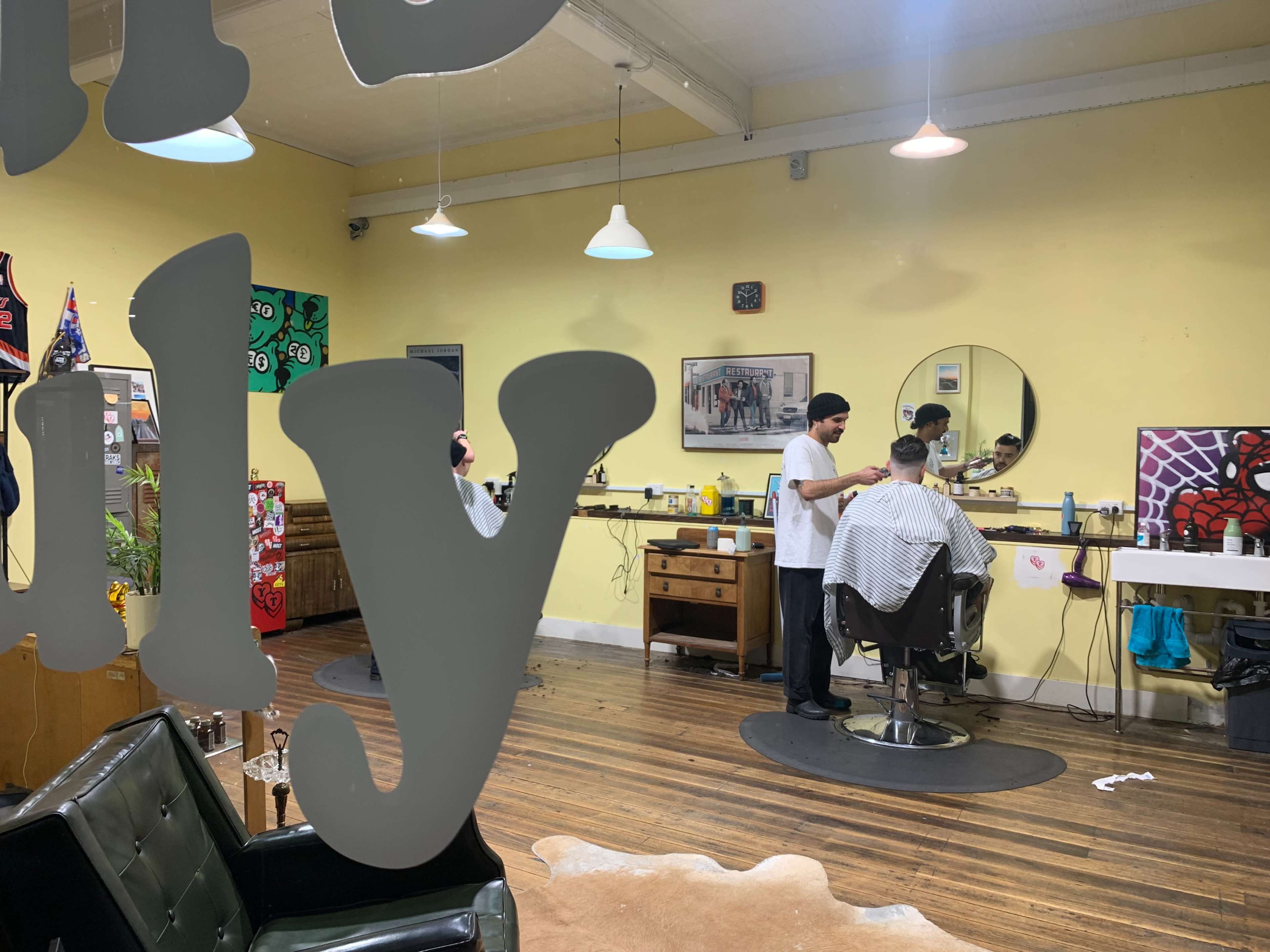 A barbershop interior with two barbers attending to clients in styling chairs, surrounded by mirrors and colorful artwork on the walls.