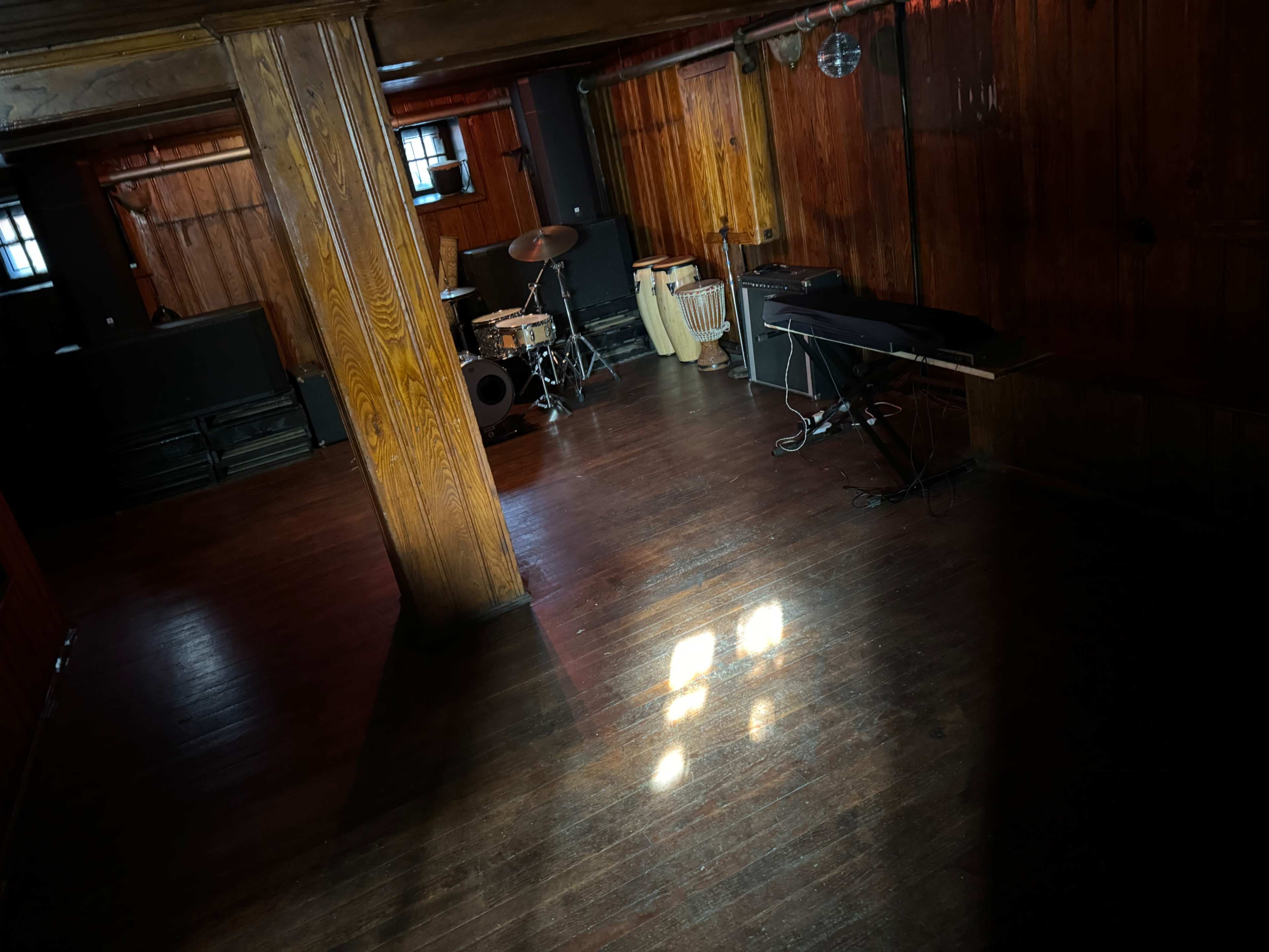 A wooden-floored room with musical instruments, including a drum set and a keyboard, positioned under low lighting.