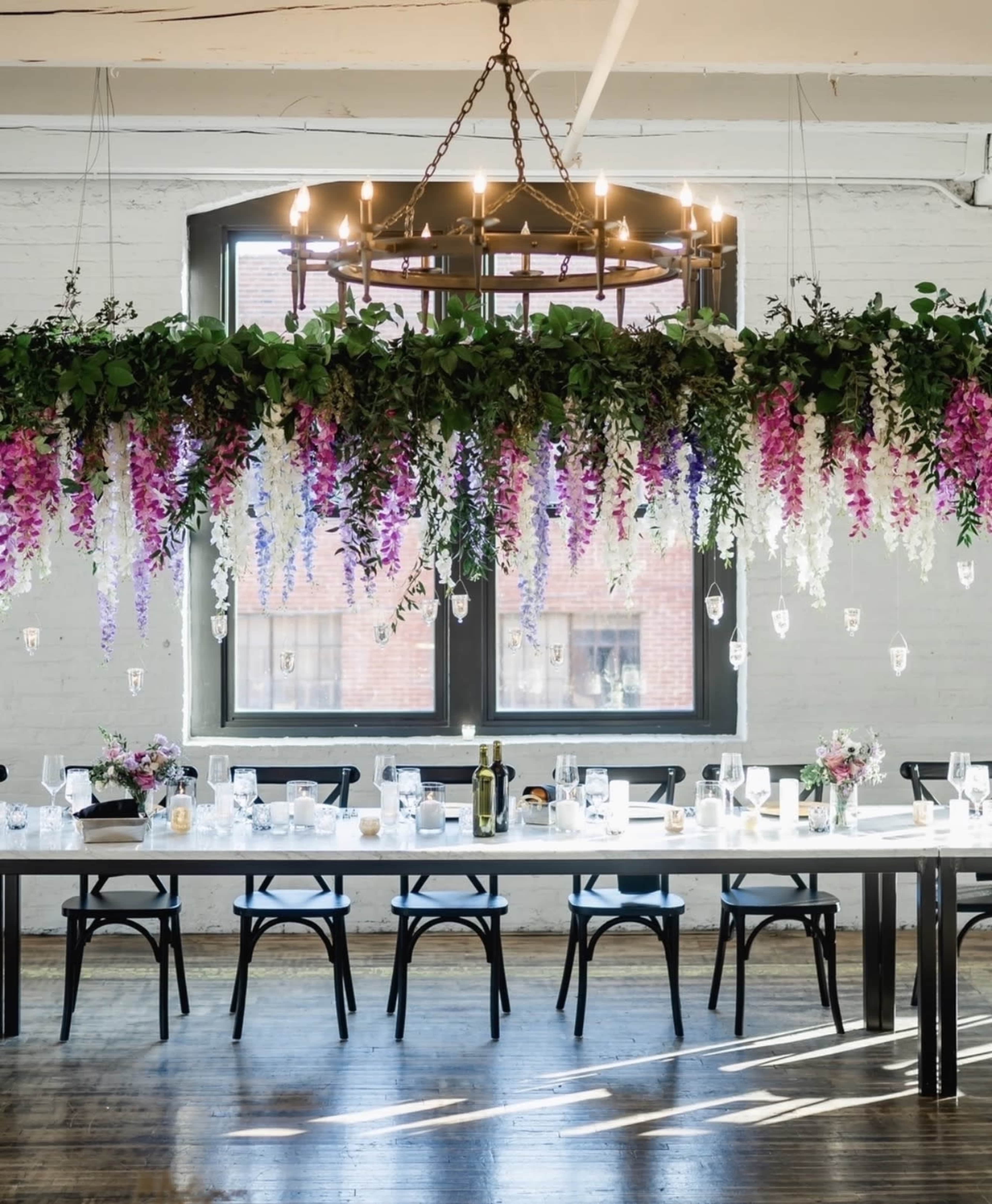 A long dining table is set under a decorative chandelier adorned with hanging floral arrangements in a bright, airy room with large windows.