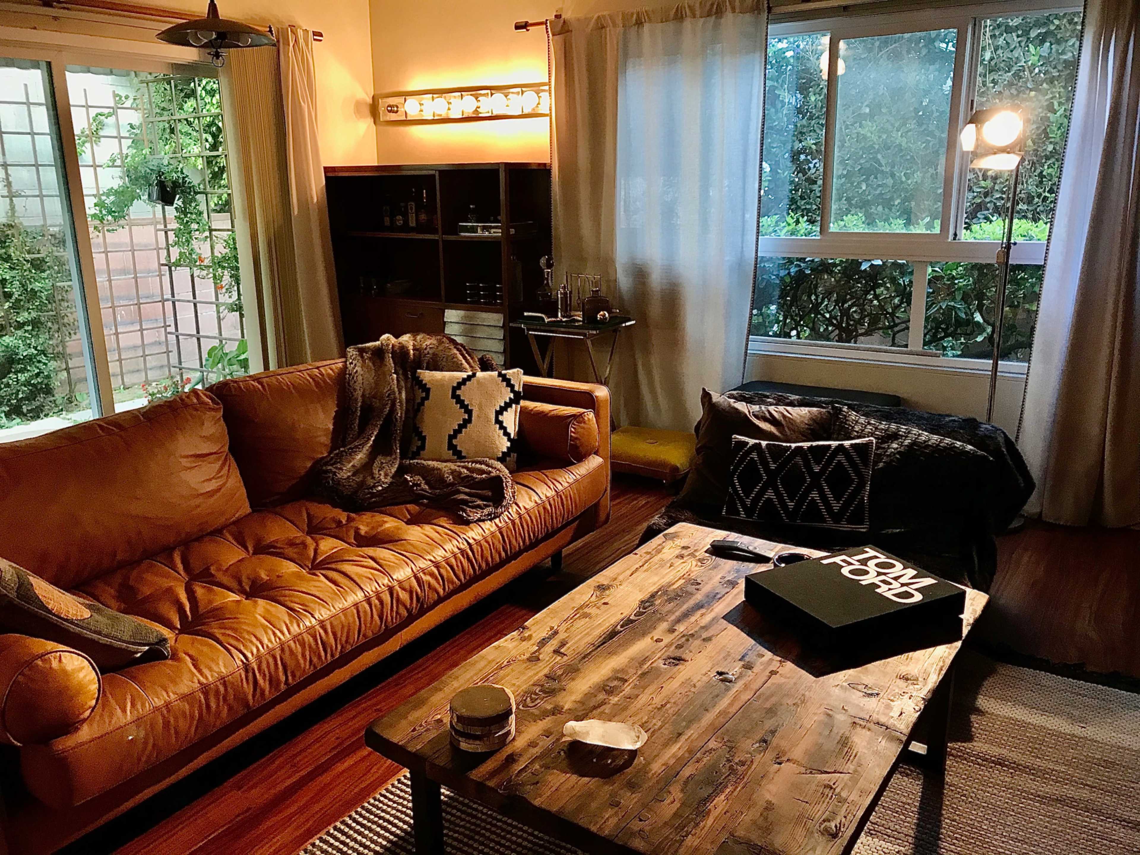 The image shows a cozy living room featuring a brown leather couch, a wooden coffee table, and large windows with sheer curtains.