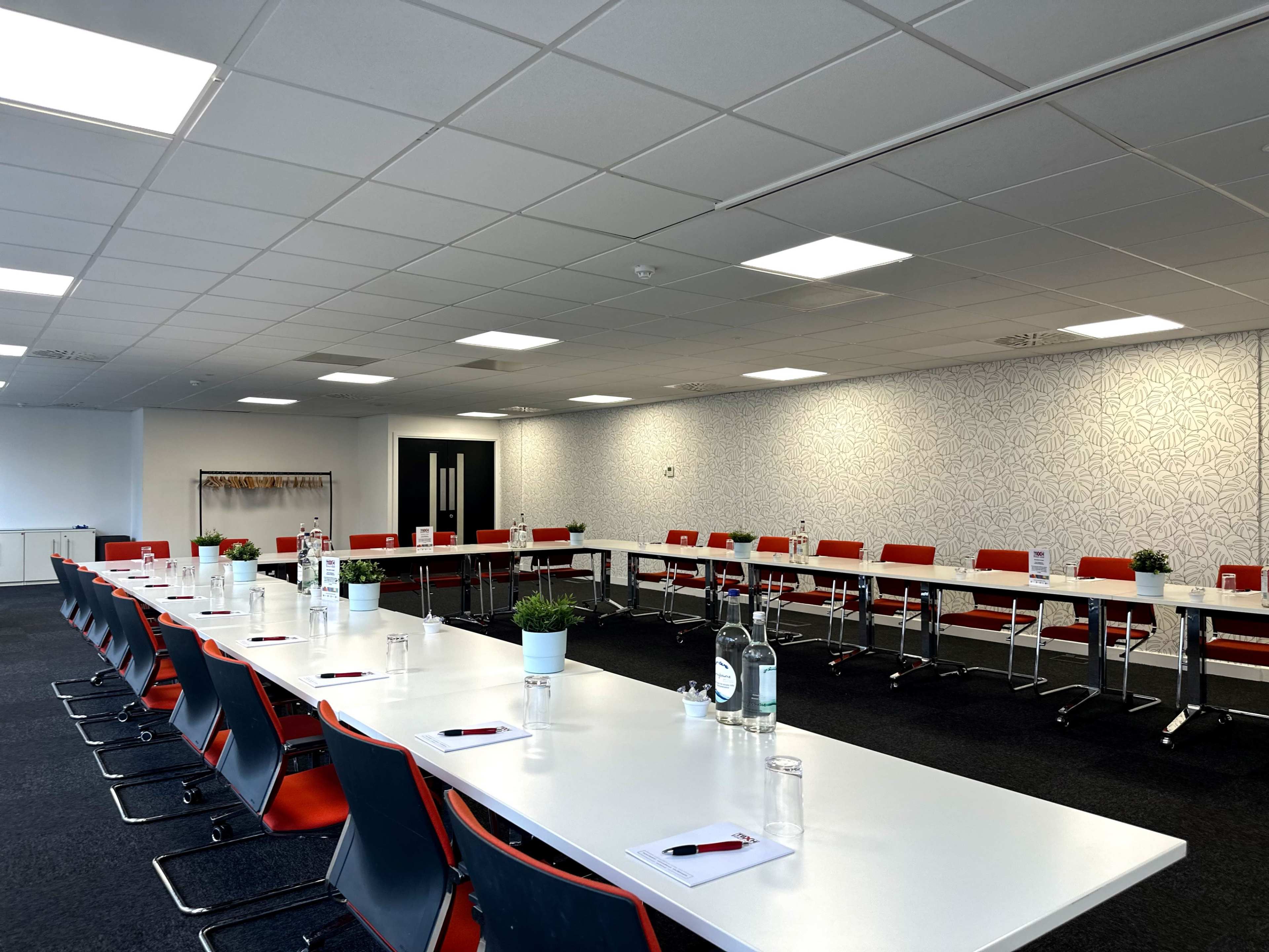 A large conference room features a long, white table surrounded by red chairs, with potted plants and water bottles on the table.