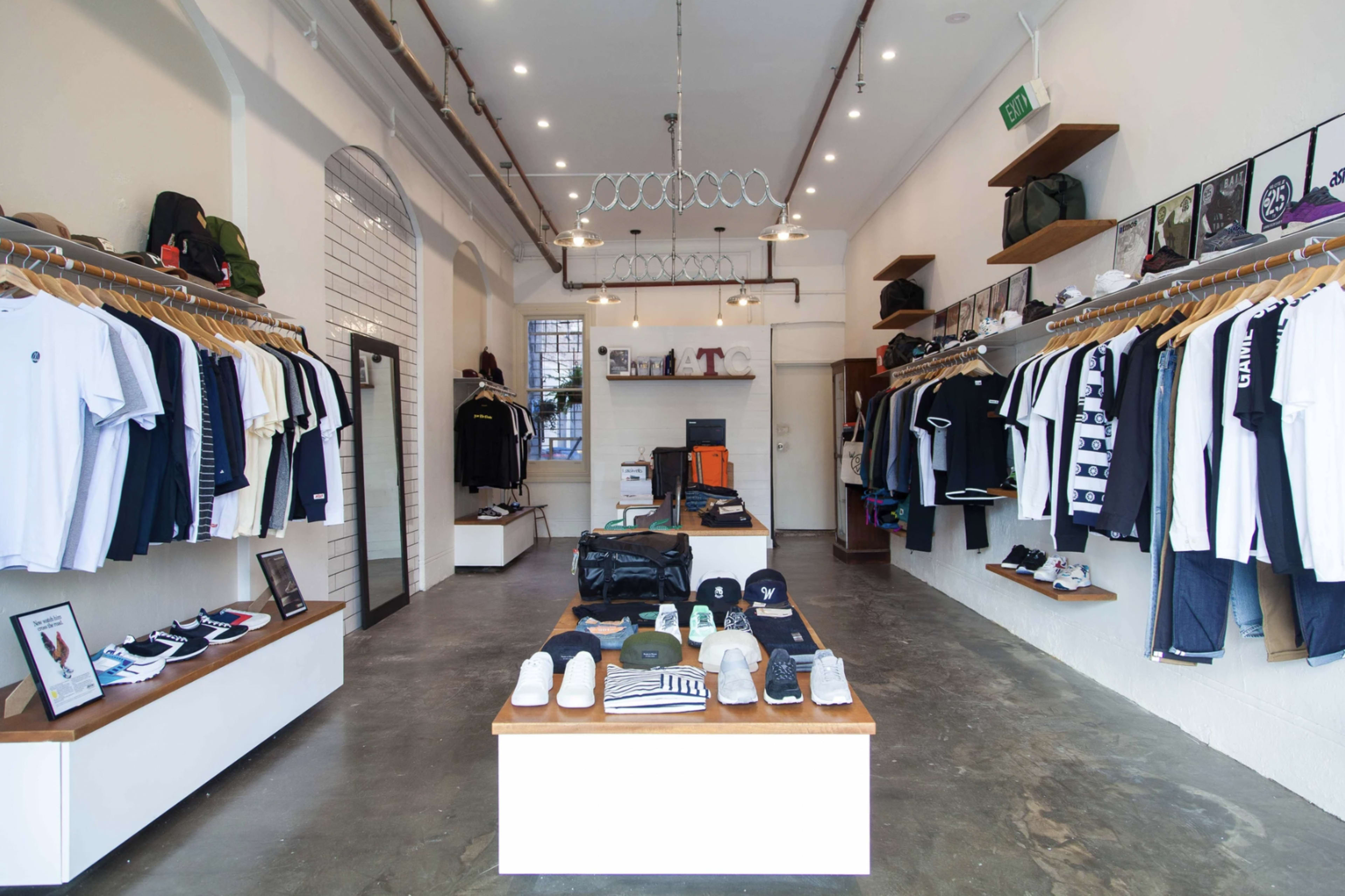 A clothing store interior with shelves displaying various garments, organized neatly along the walls, and a central table featuring shoes and accessories.