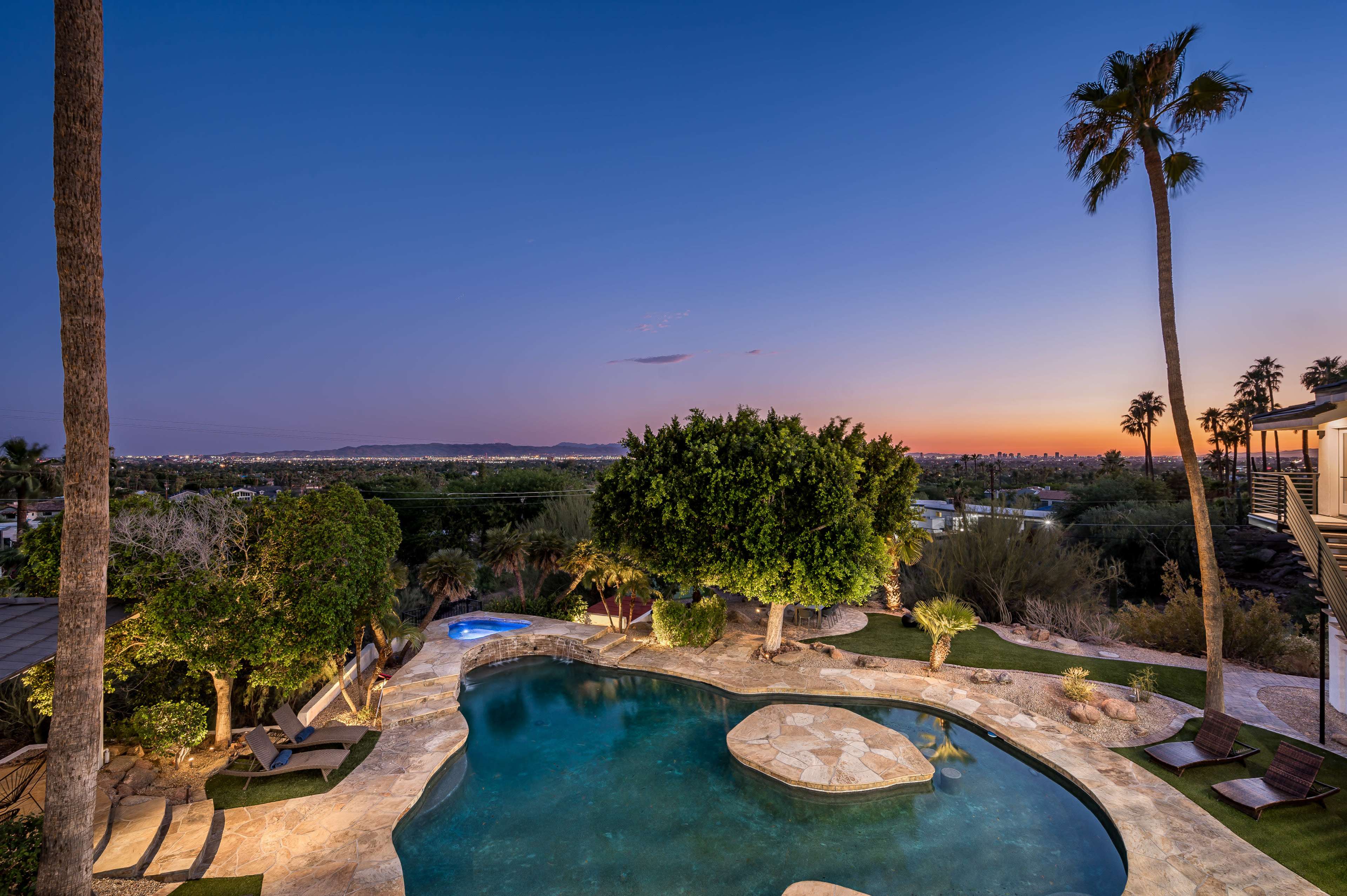 A backyard pool area features a rock formation and lush landscaping, with palm trees silhouetted against a twilight sky.