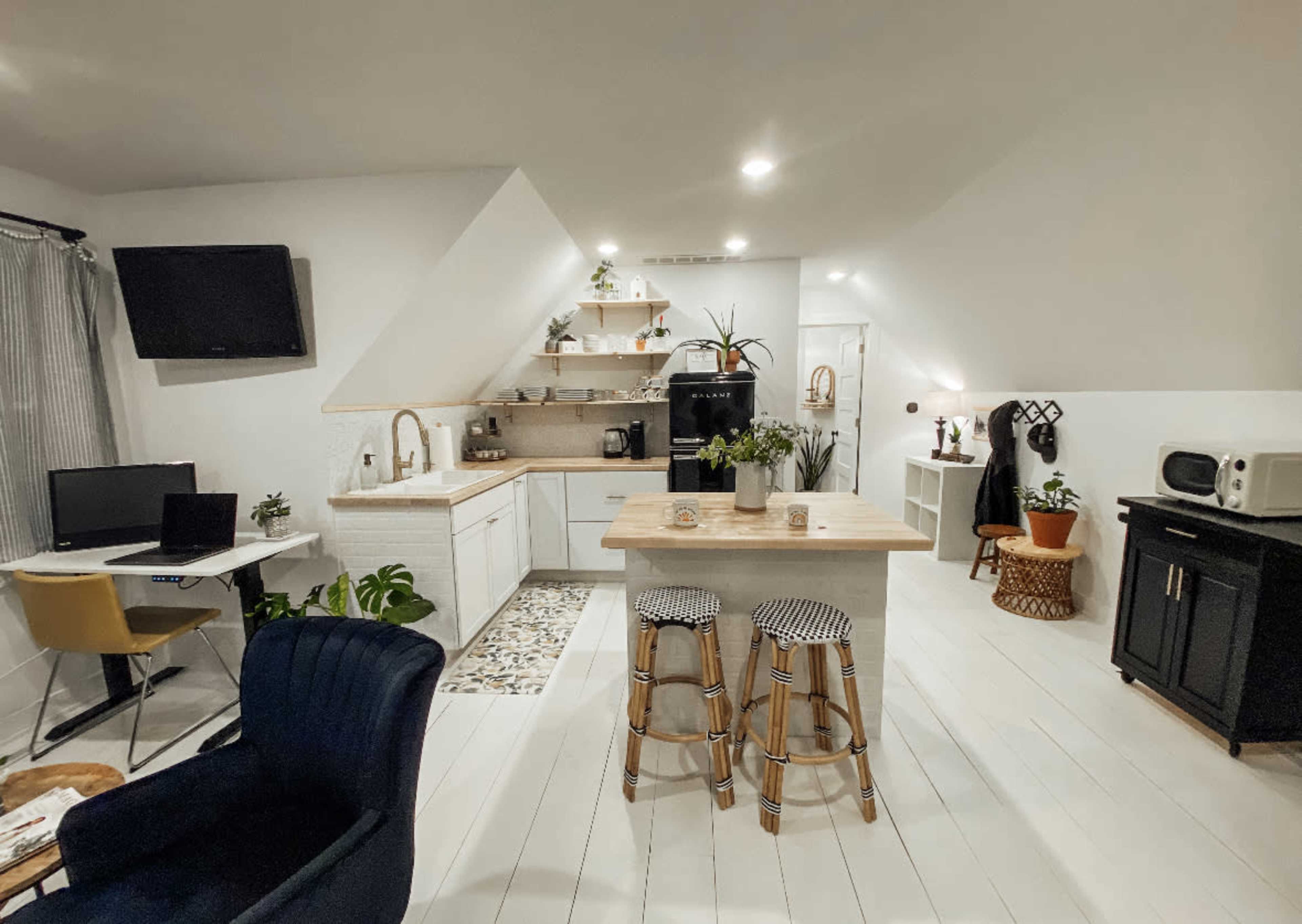 A modern, minimalist kitchen and living area with white walls, a wooden island, and two bar stools, alongside a small workspace featuring a desk and chair.