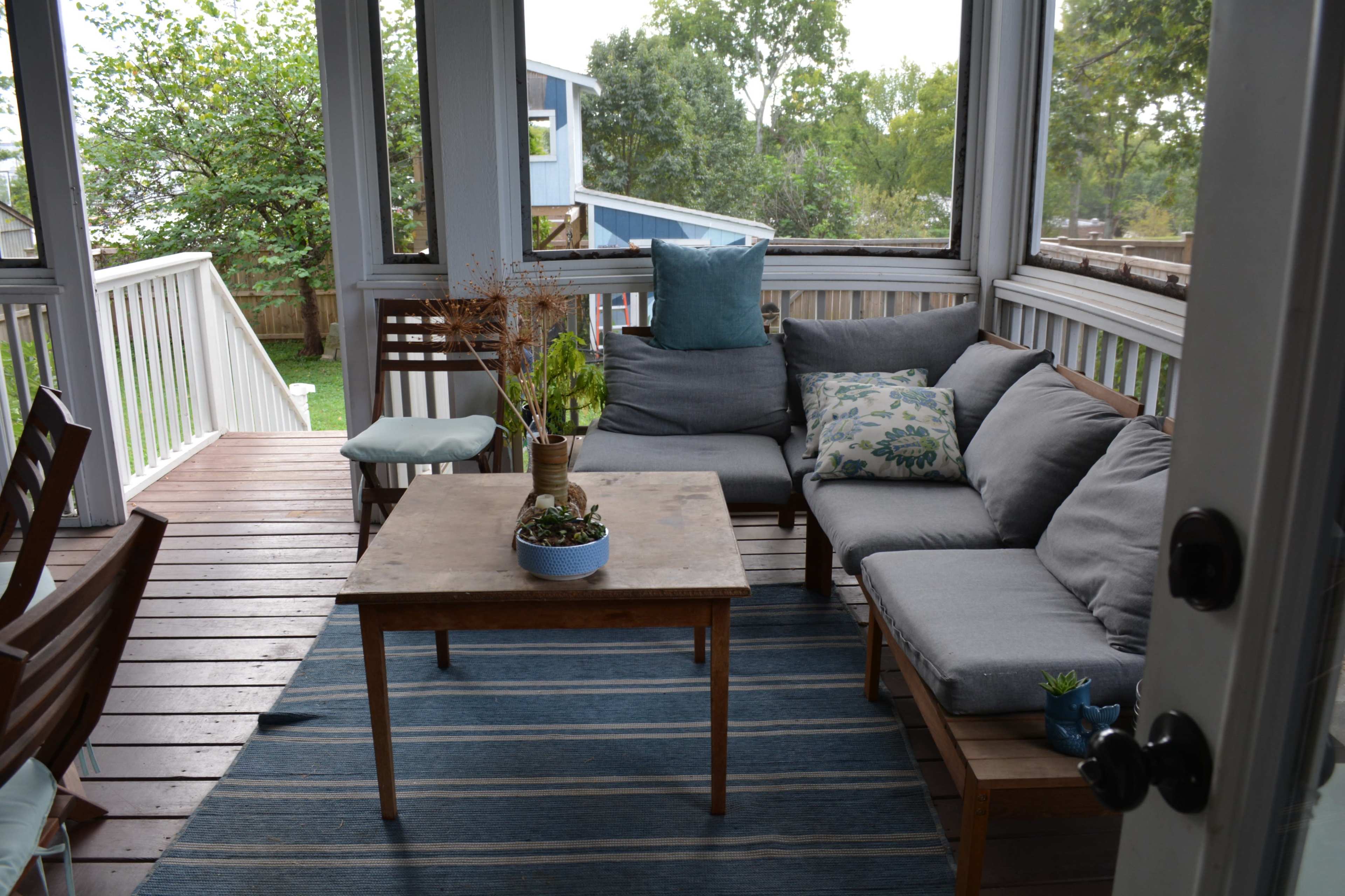 A screened-in porch features a gray sectional sofa arranged around a wooden coffee table, with potted plants and decorative cushions, set against a backdrop of greenery.