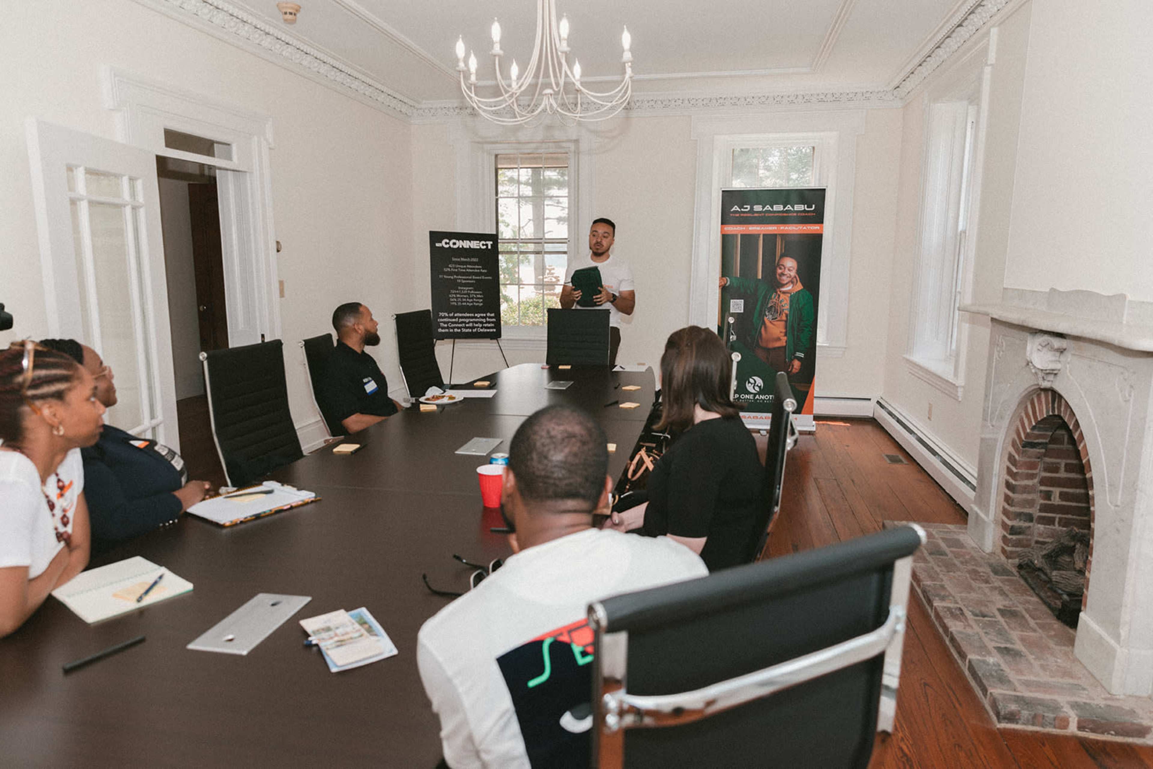 A group of people sits around a conference table while a speaker stands at the head of the table presenting.