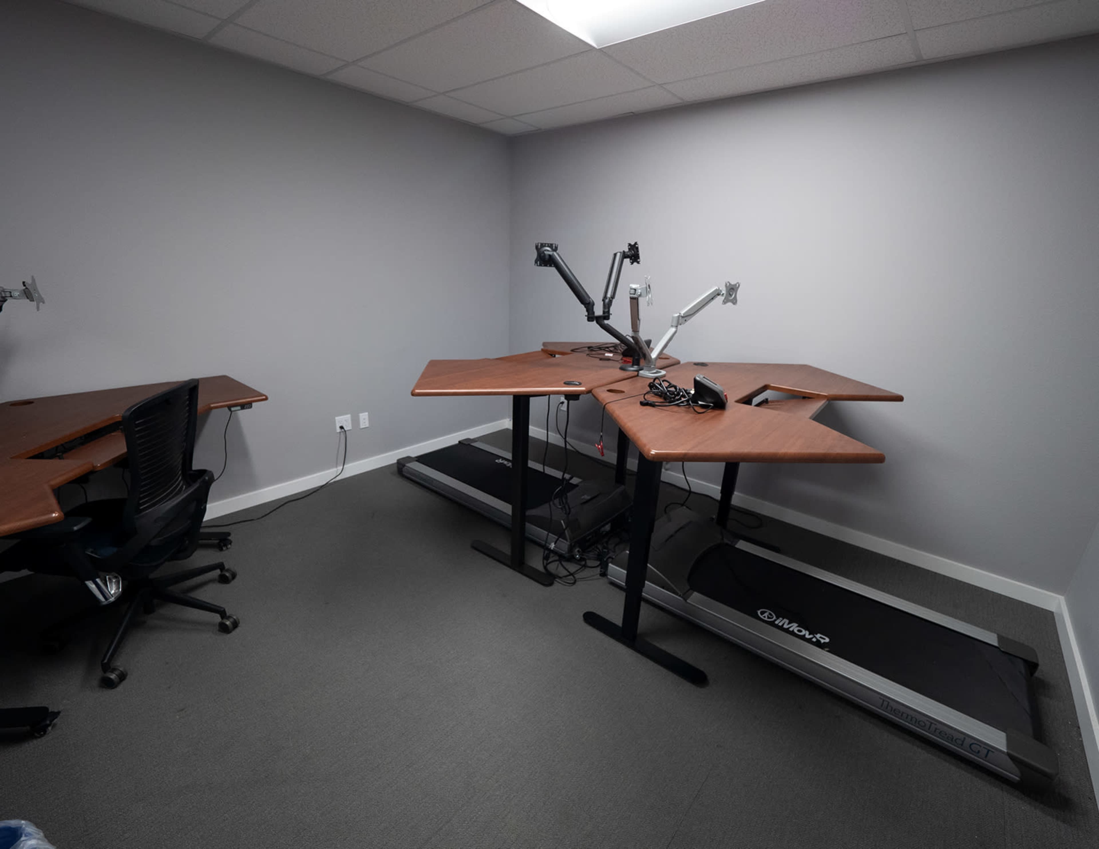 A minimalist office space featuring two standing desks, one with a treadmill, against a plain gray wall.