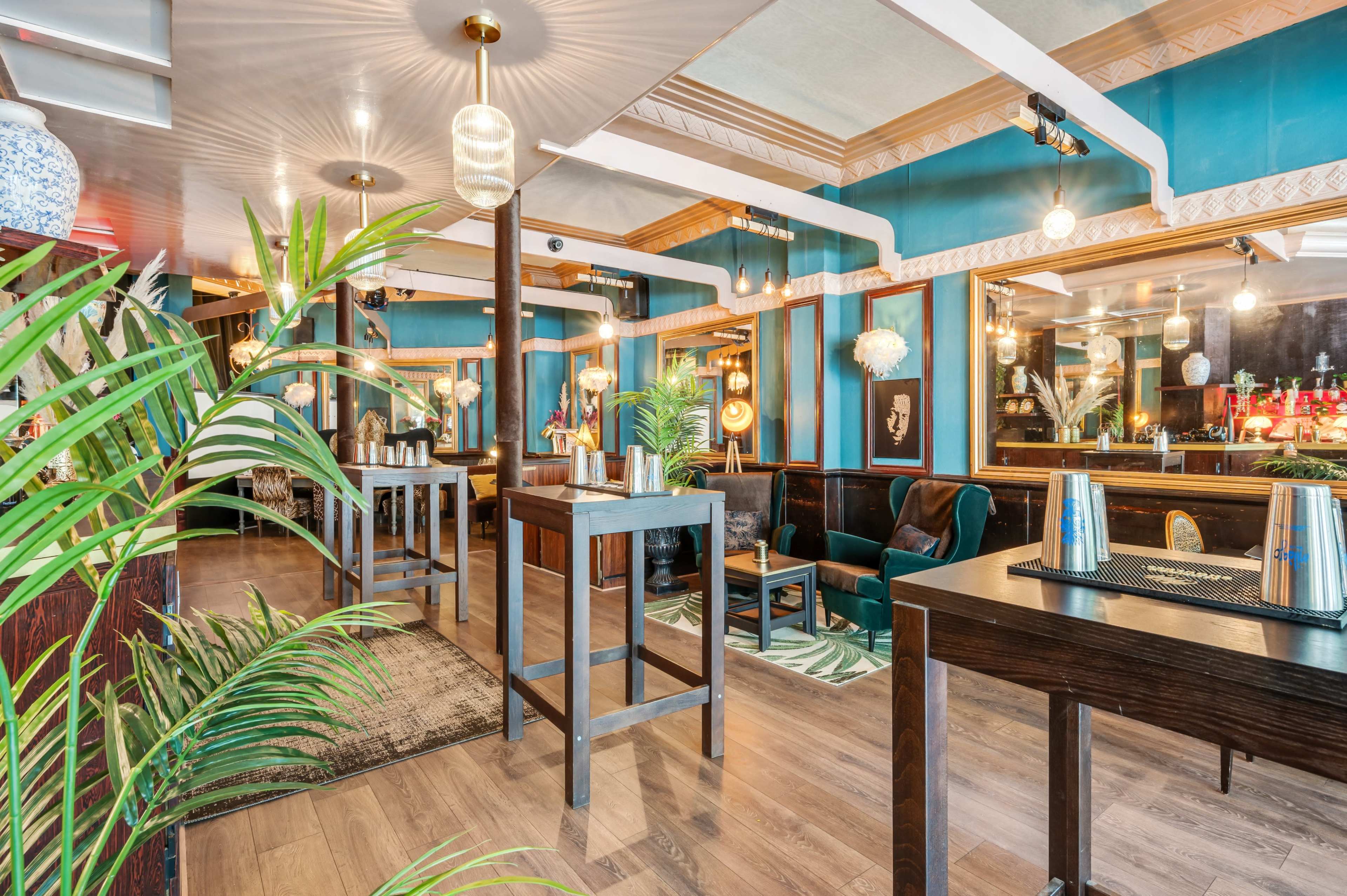 A stylish bar interior featuring wooden tables, plants, mirrors, and a seating area with green upholstery.