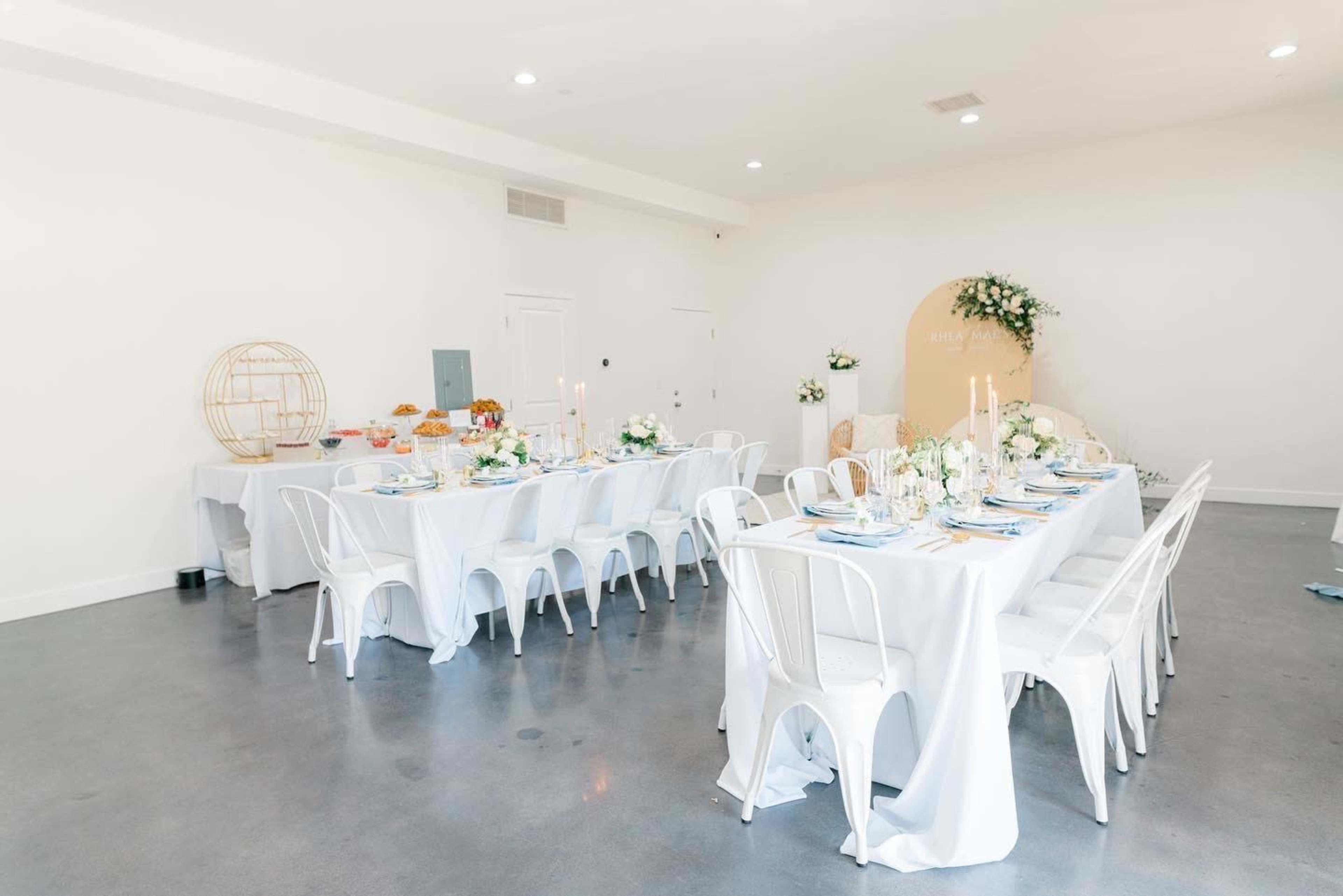 The image shows a light-filled event space with two long tables covered in white tablecloths, decorated with floral arrangements and candles.