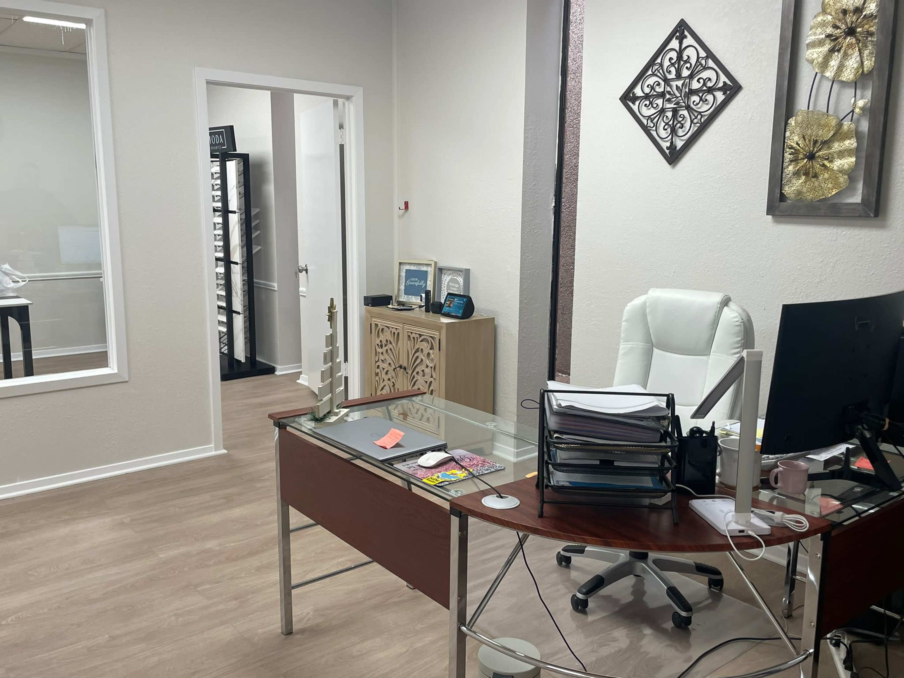 An office space featuring a glass desk, a computer monitor, a white office chair, and decorative wall art.