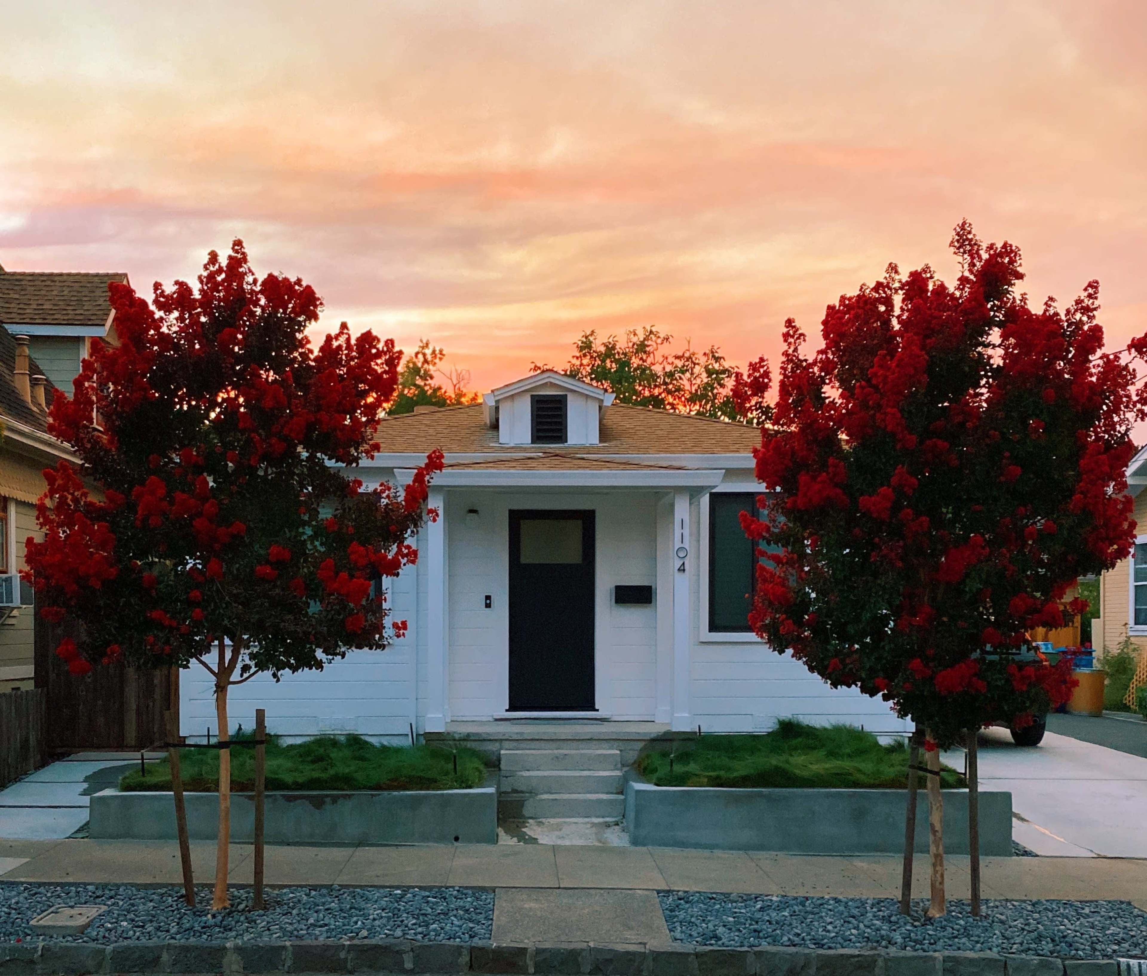 A single-story white house with two red flowering trees in the front yard is framed by a colorful sunset sky.