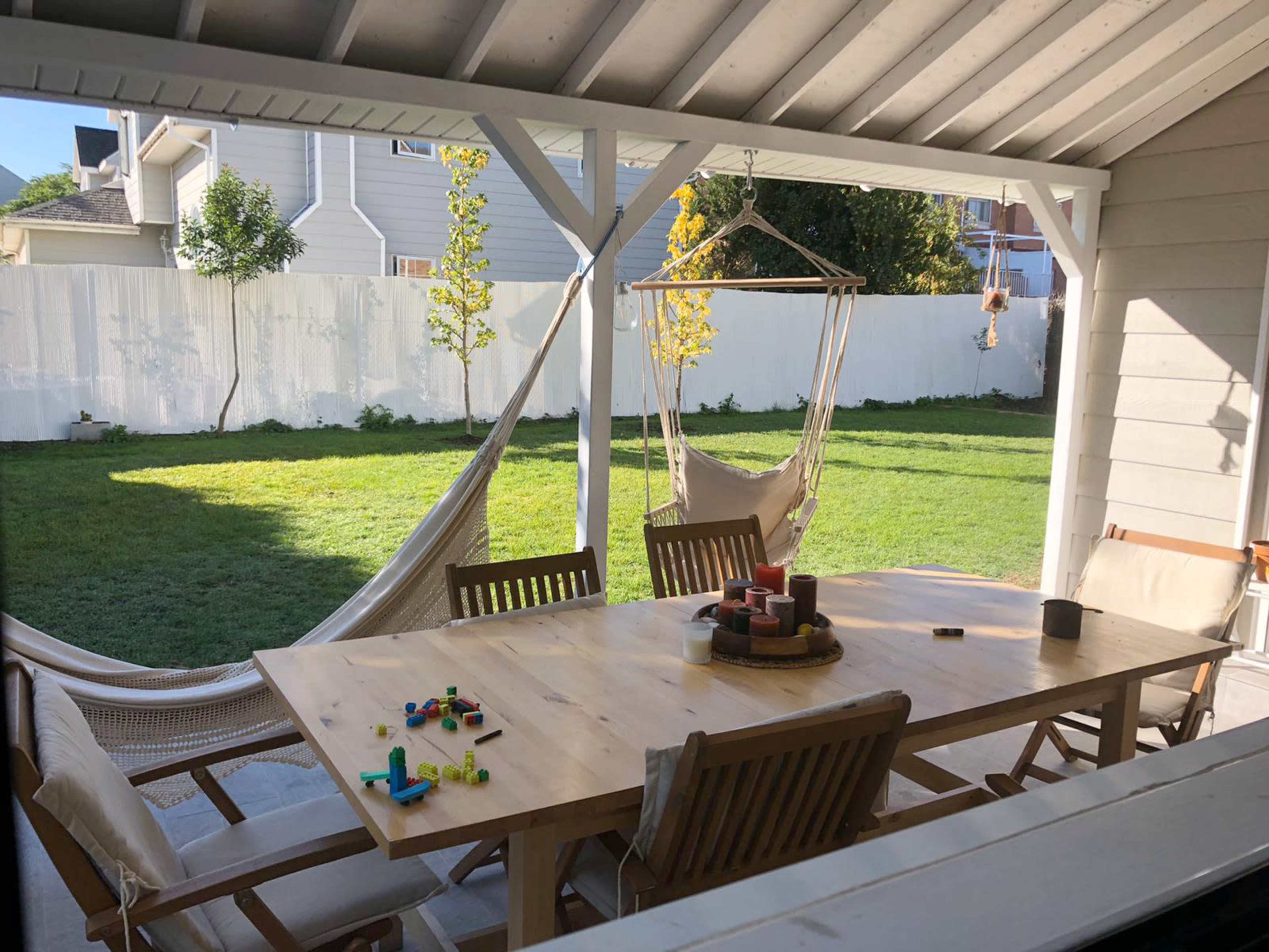 The image shows a patio area with a large wooden dining table surrounded by chairs, a hammock hanging in the background, and a green lawn beyond a white fence.