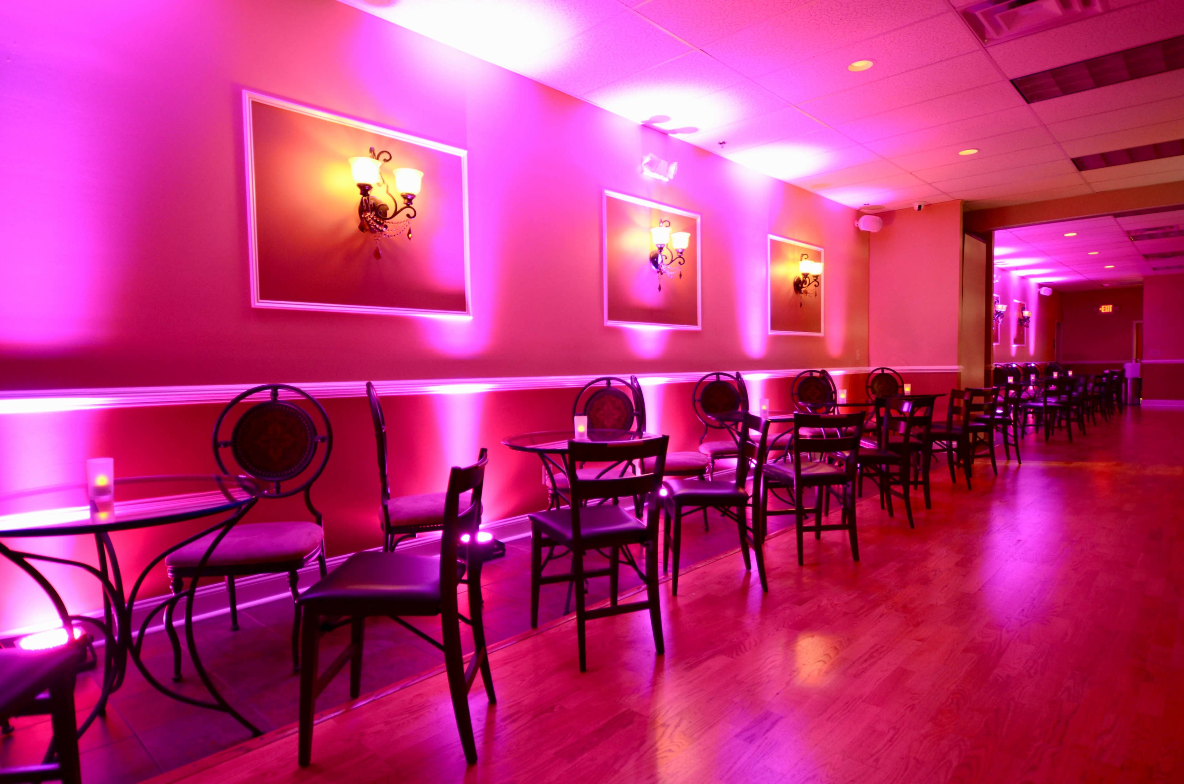 A narrow, well-lit lounge with purple-hued walls and chairs arranged around small tables.