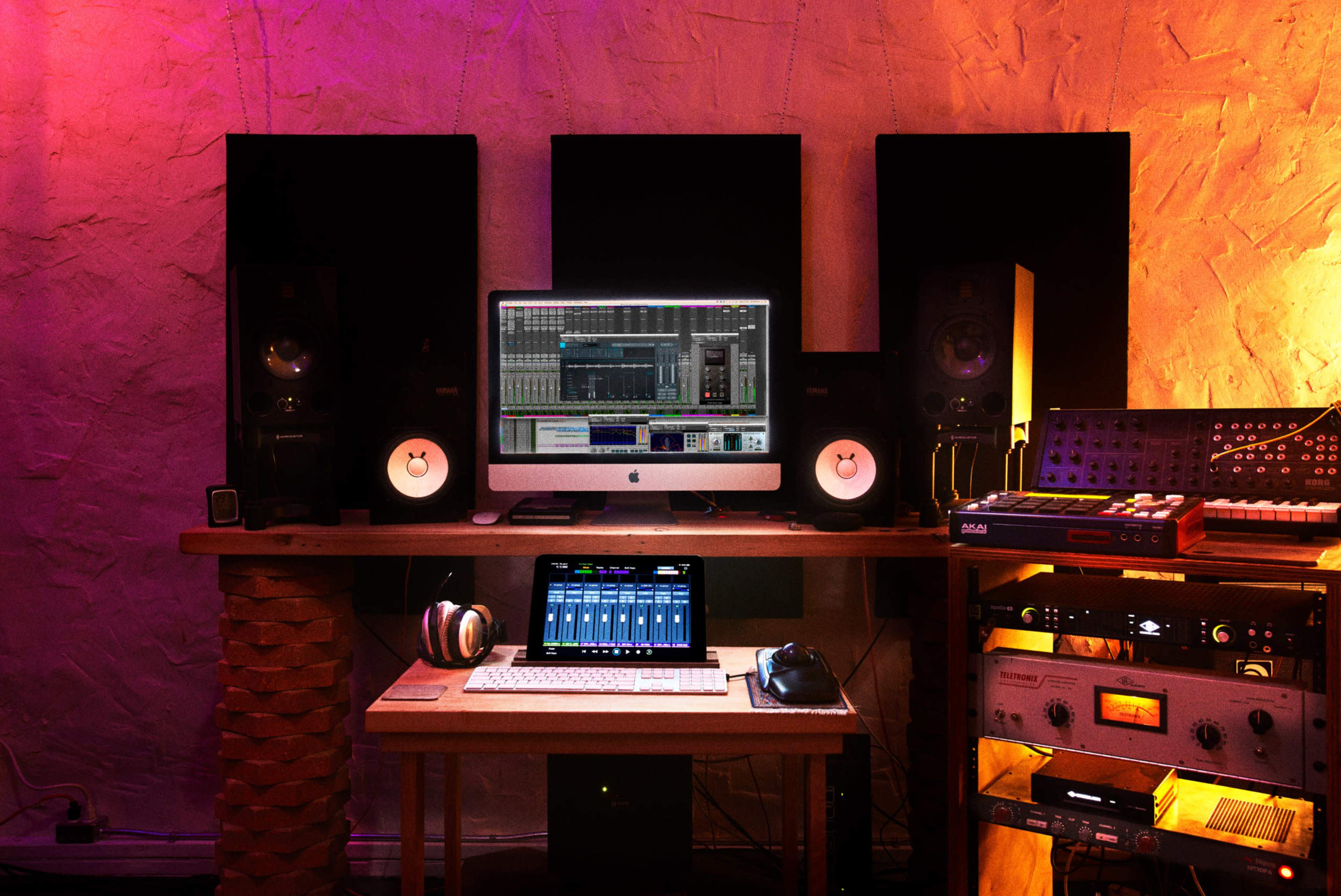 A music production setup featuring a computer monitor, audio speakers, a keyboard, headphones, and various electronic instruments on a wooden desk against a textured wall.