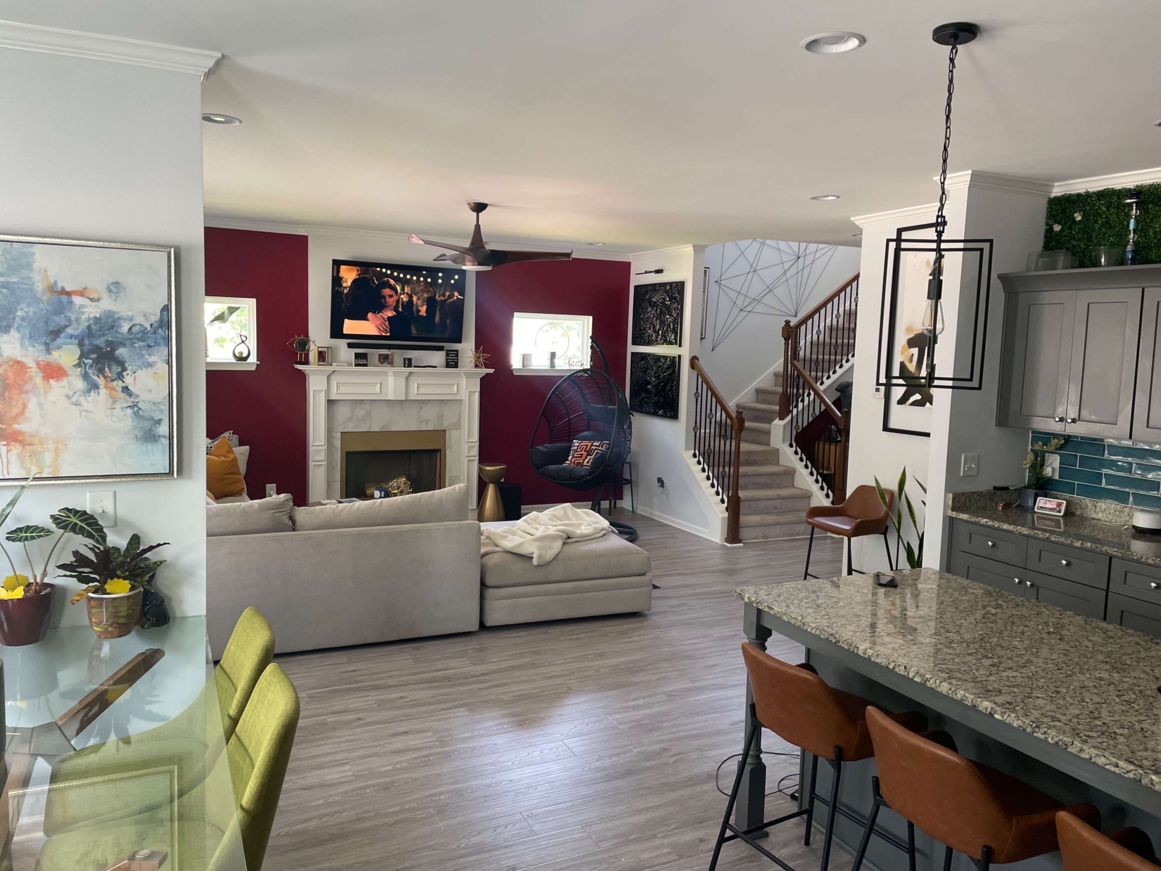 The image shows a modern, open-concept living space featuring a gray sofa, a hanging chair, a television mounted above a fireplace, and a kitchen island with bar stools.