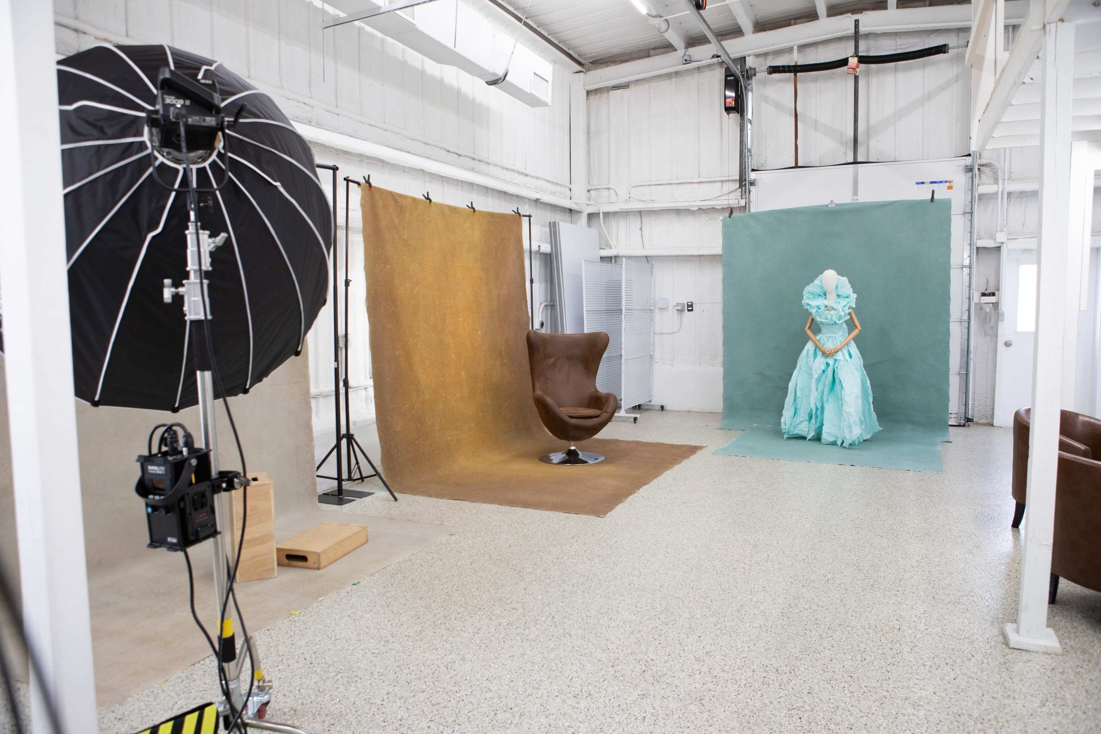 The image displays a photography studio featuring two backdrops—one brown and one green—along with a large black umbrella softbox and a mannequin dressed in a turquoise dress.